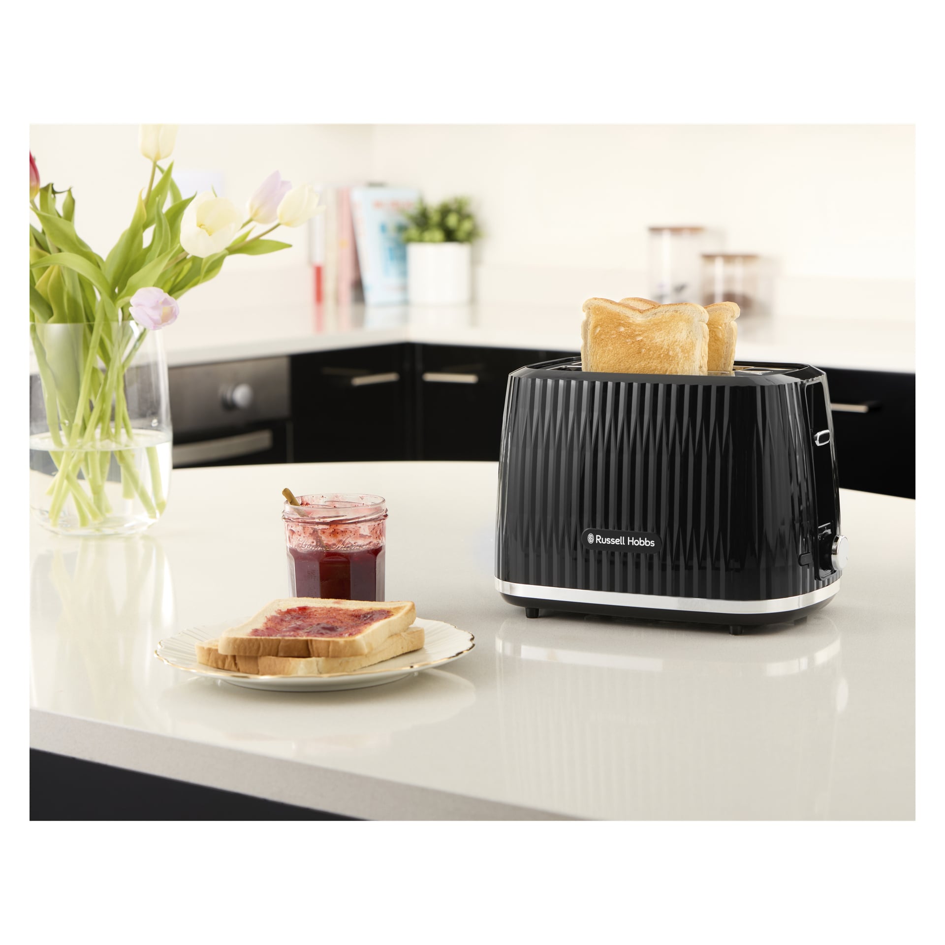 toster Russell Hobbs z dwoma plastami stoi na blacie obok talerza z tostami, czerwonym dżemem, słoikiem na dżem i wazonem z kwiatami.