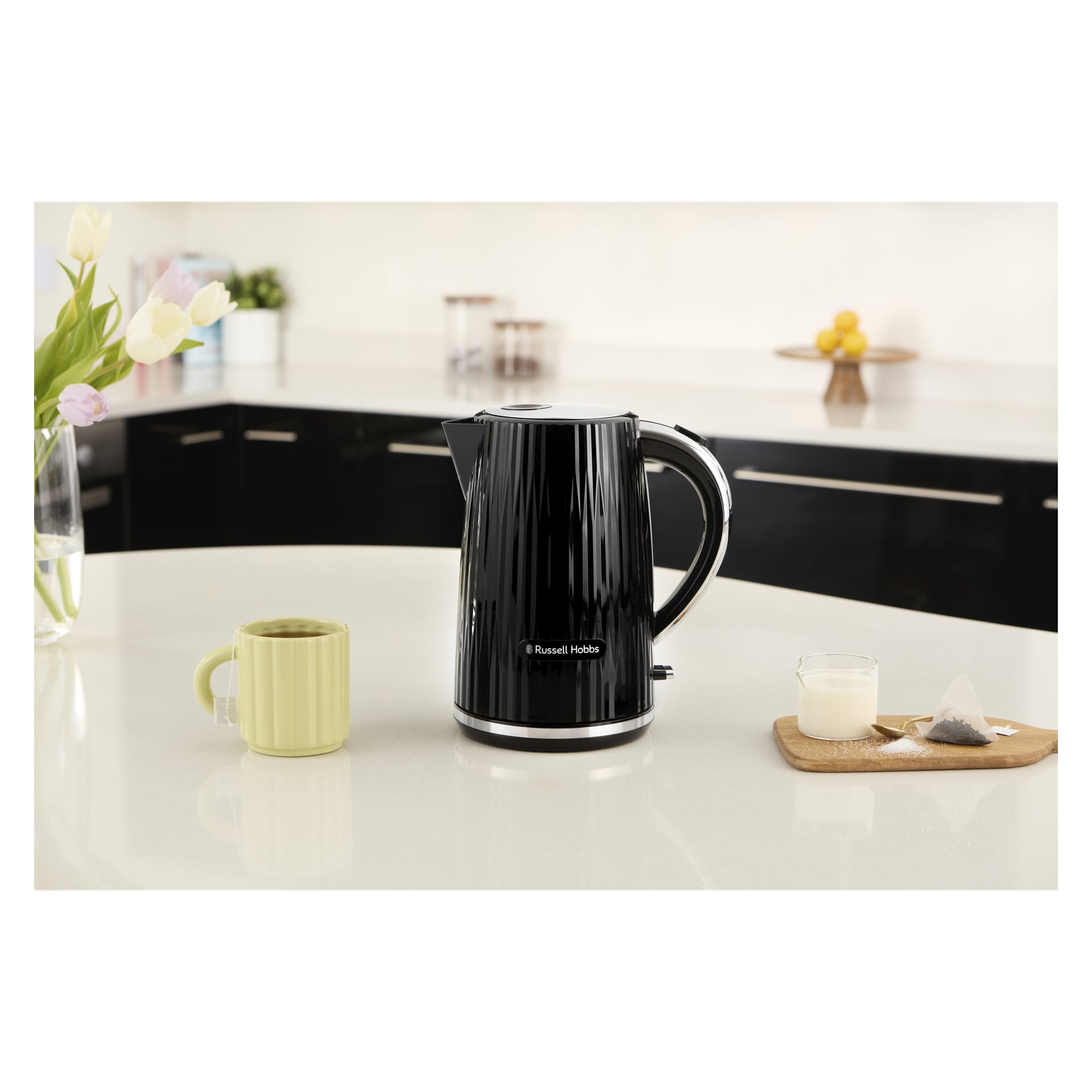 En svart Russell Hobbs Eden-kjele på en benkeplate ved siden av en grønn kopp, melkeglass, tepose, trebrett med løs te og blomster.