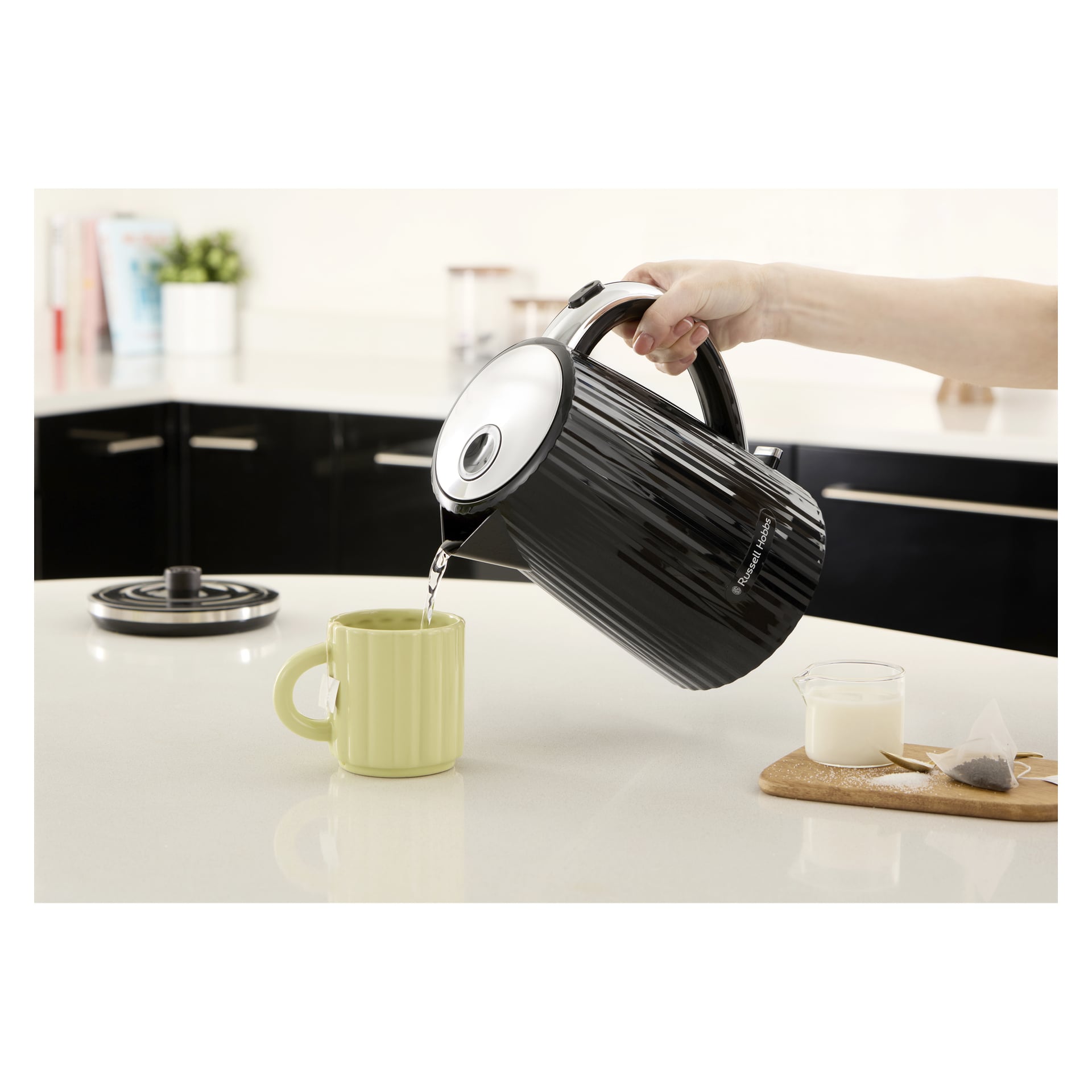 Varmt vann helles fra en svart Russell Hobbs Kettle Eden i en lysegrønn kopp; I nærheten er det en skje, en tepose og en kopp melk.