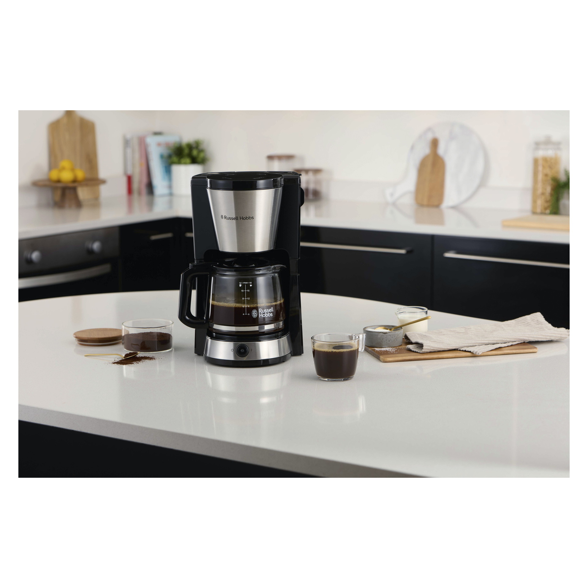 Ekspres do kawy Russell Hobbs Heaton z szklaną karafką na blacie obok dwóch filiżanek do kawy, mielonej kawy, cukru i serwetki.