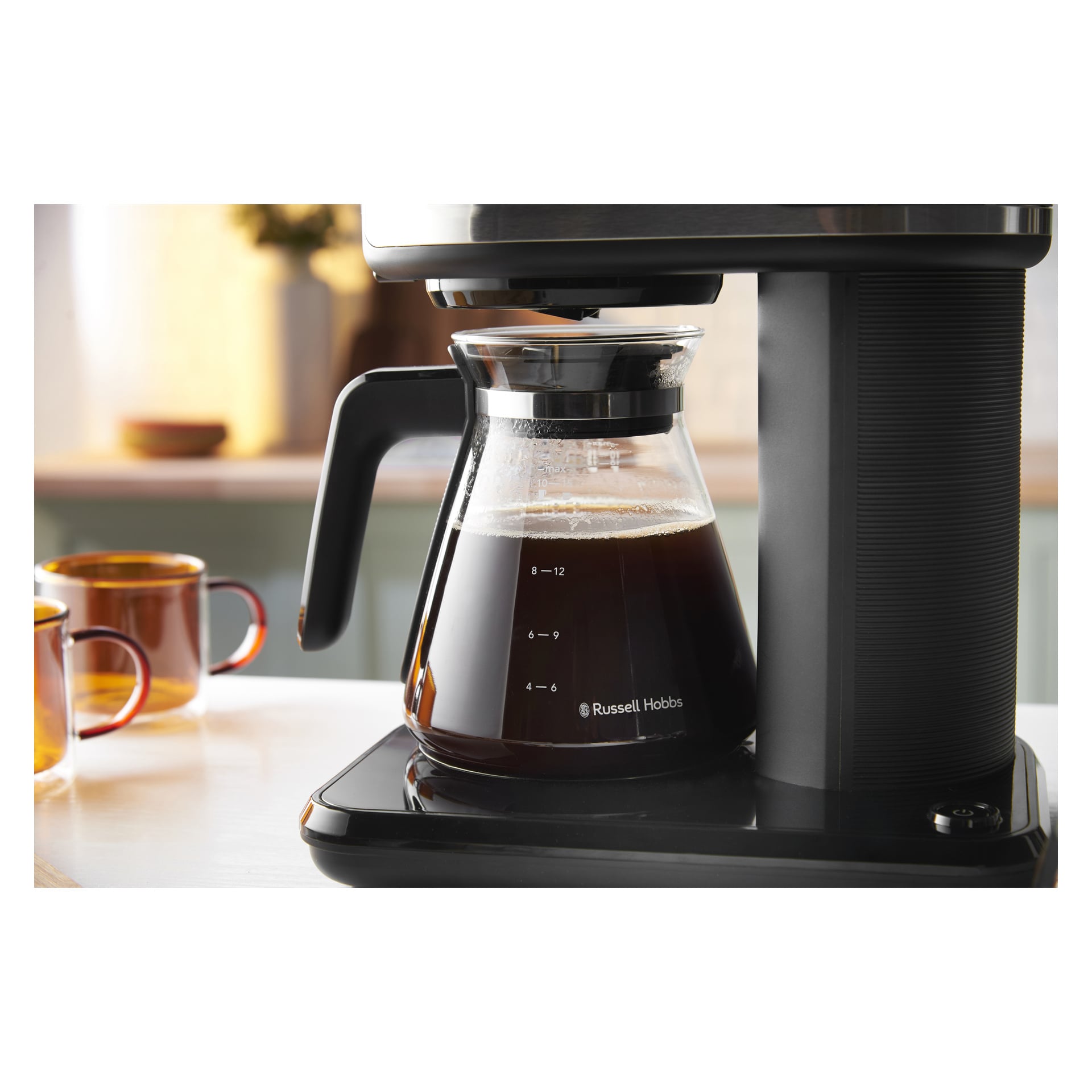 En glaskaffekanna fylld med bryggt kaffe står på en Russell Hobbs Attentiv Black-maskin; Två bruna muggar står bakom på disken.