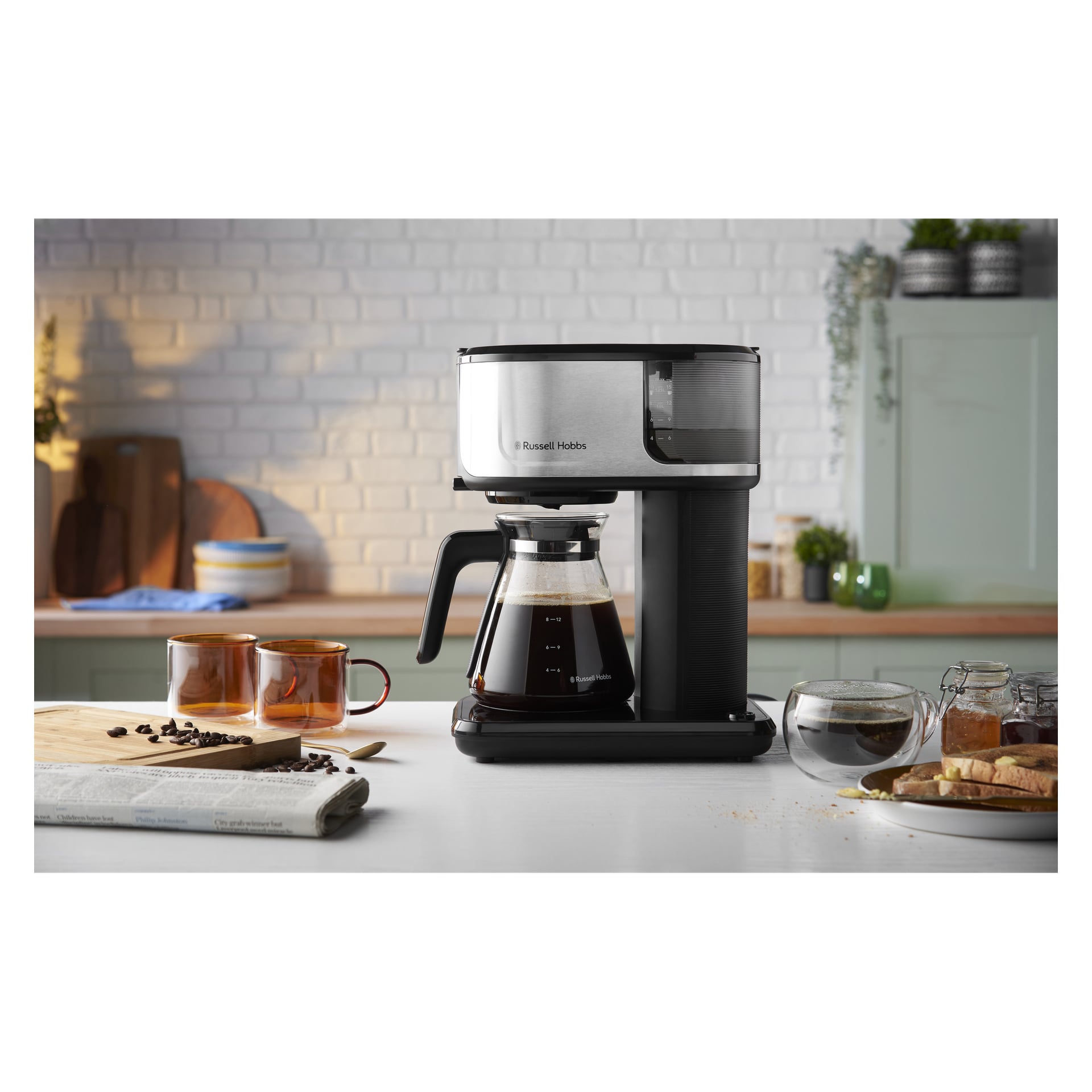 En Russell Hobbs Attentiv Black kaffebryggare på köksbänken med muggar, bröd, vattenskål, kaffebönor och tidning.