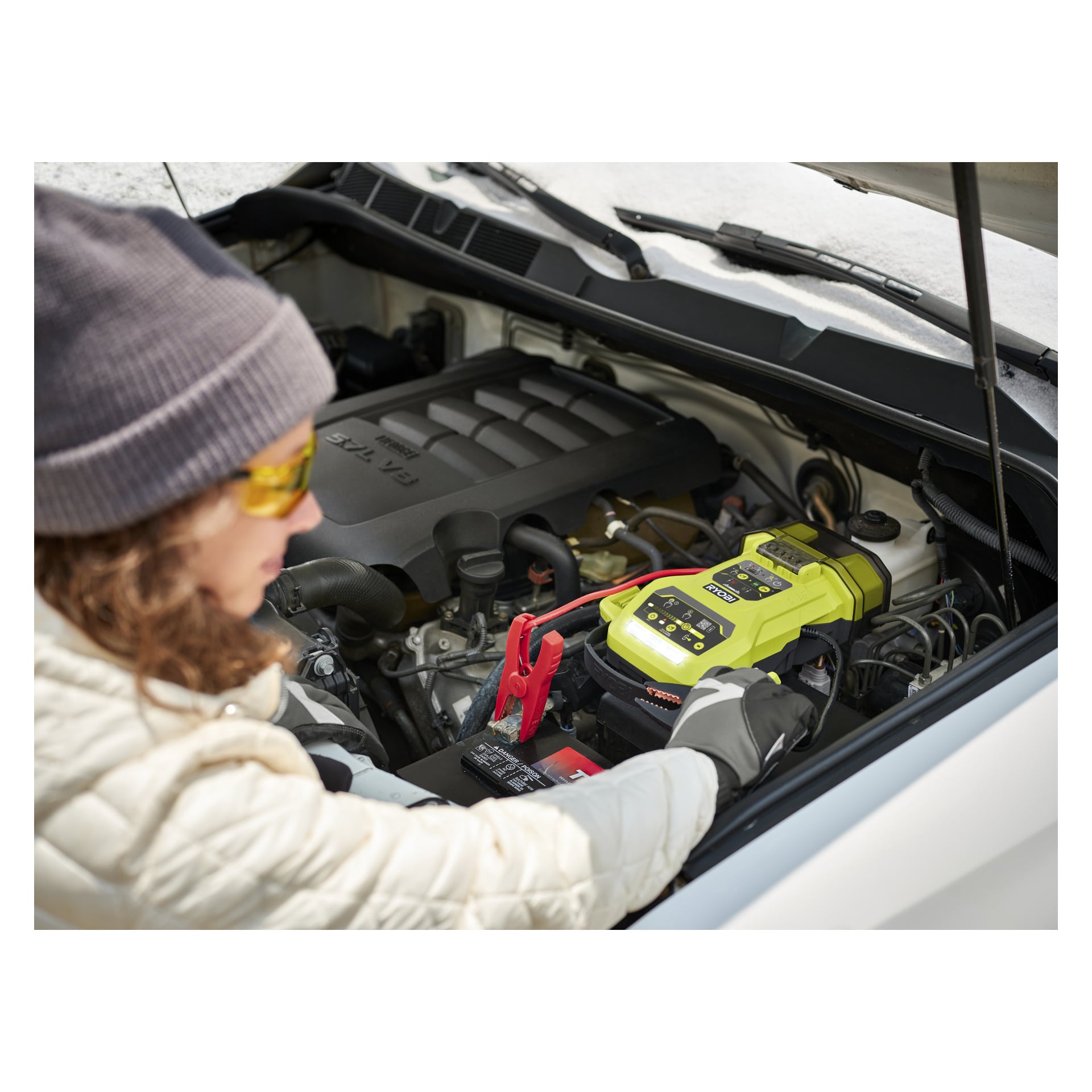 Med grå hatt och gula glasögon använder personen en Ryobi One+ Jump Starter RJSC18-0 för att starta ett bilbatteri.
