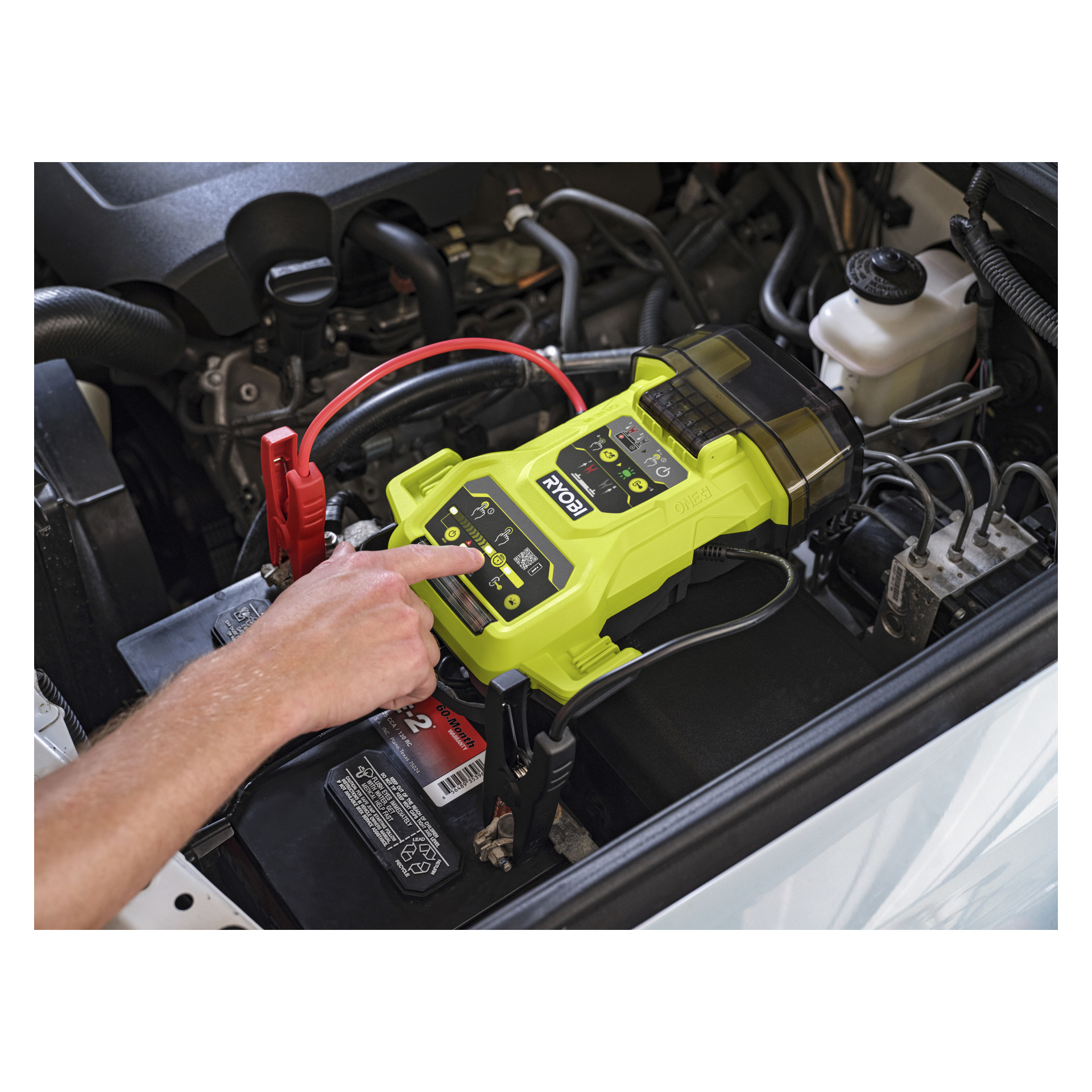 Någon använder en Ryobi One+ Jump Starter RJSC18-0 och trycker på en knapp när kablarna kopplas till ett bilbatteri under den öppna huven.