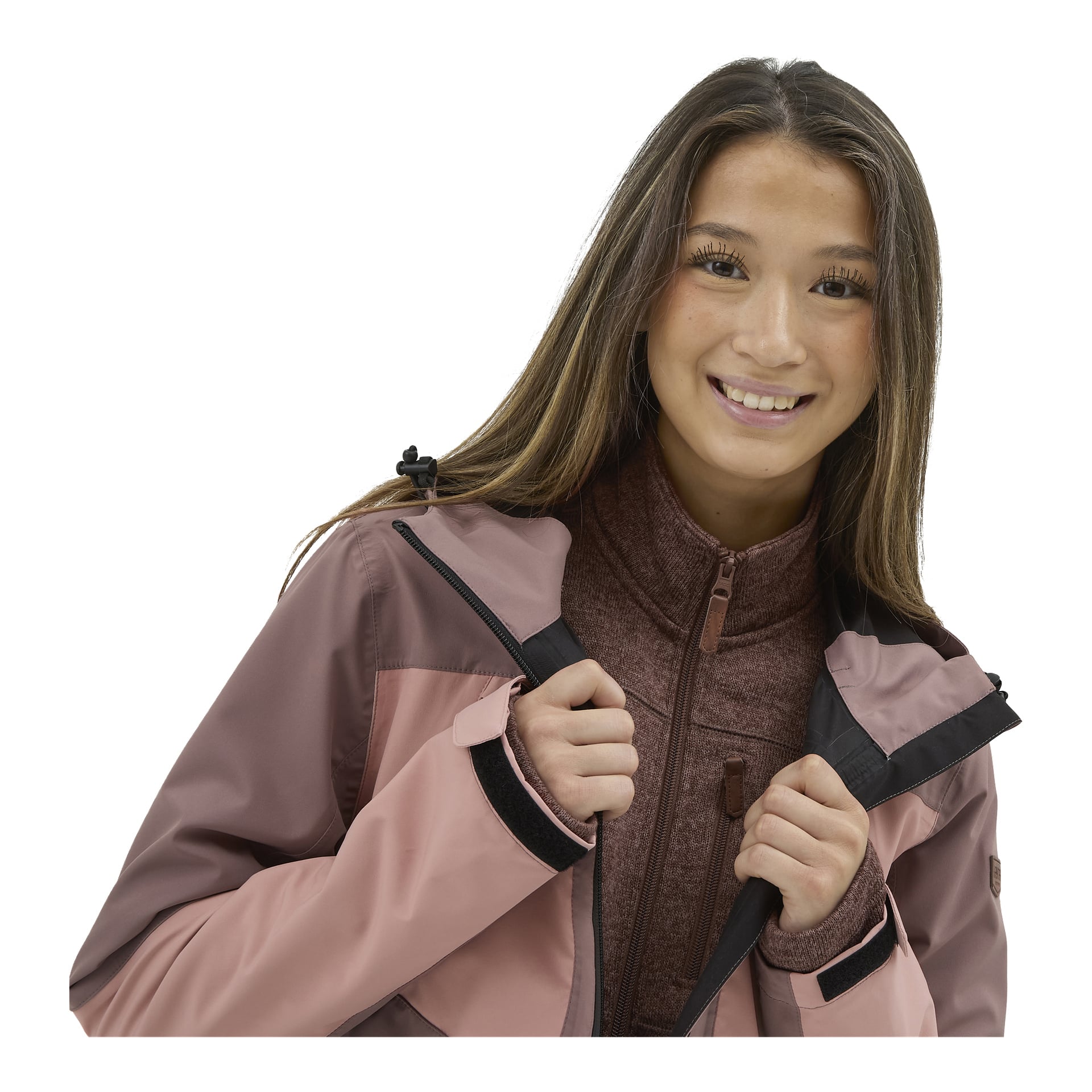En ung kvinna med långt brunt hår ler mot kameran medan hon justerar kragen på sin damstickade fleecetröja "MALÅ by Kayoba Outdoor", som bärs över en zip-up-topp.