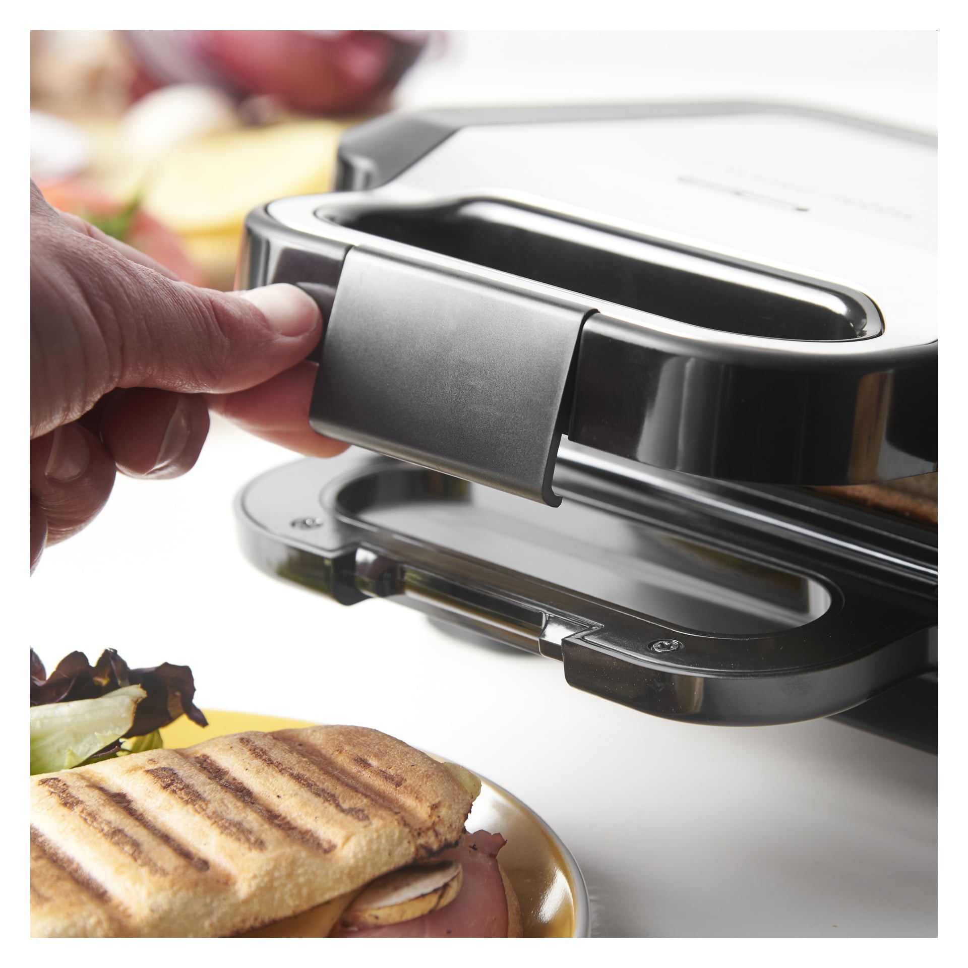 Käsi nostaa suljetun Russell Hobbs 3-in-1 Sandwich Grillin kahvaa, jonka edessä on lautasella paahtettu voileipä.