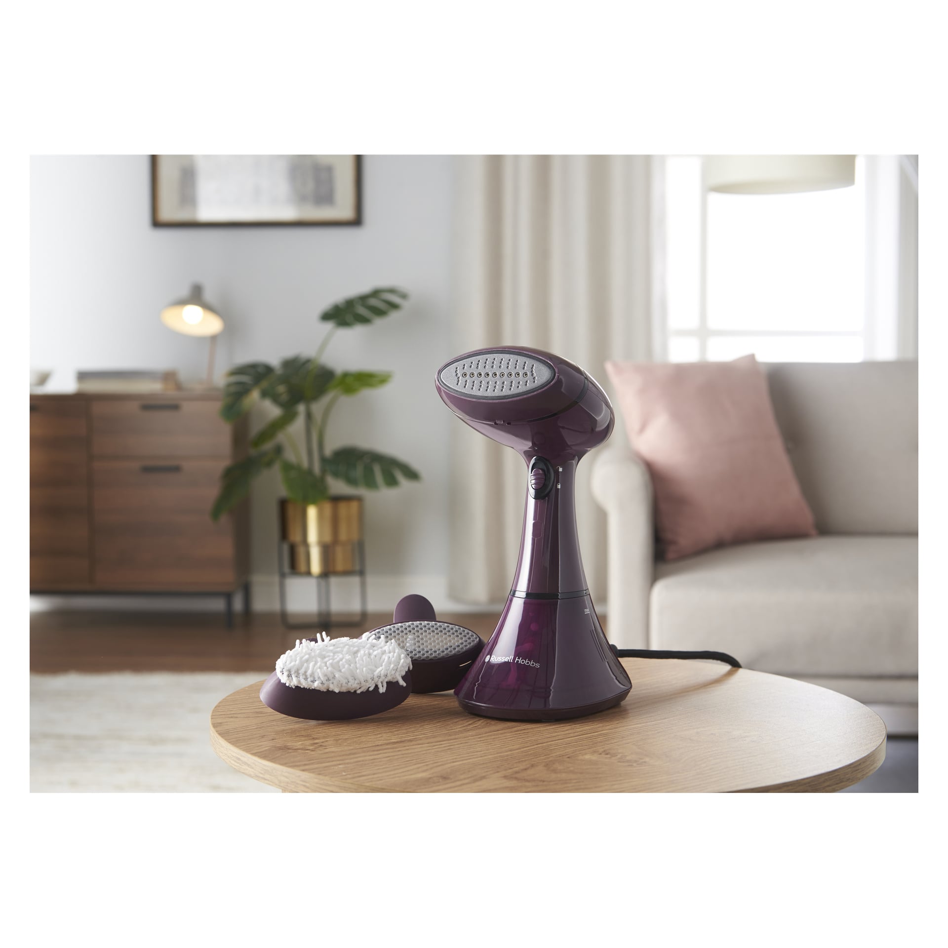 En lilla Russell Hobbs Steamer Genie Color Control står på et salongbord ved siden av mopphodet, med sofa, plante og lampe bak.