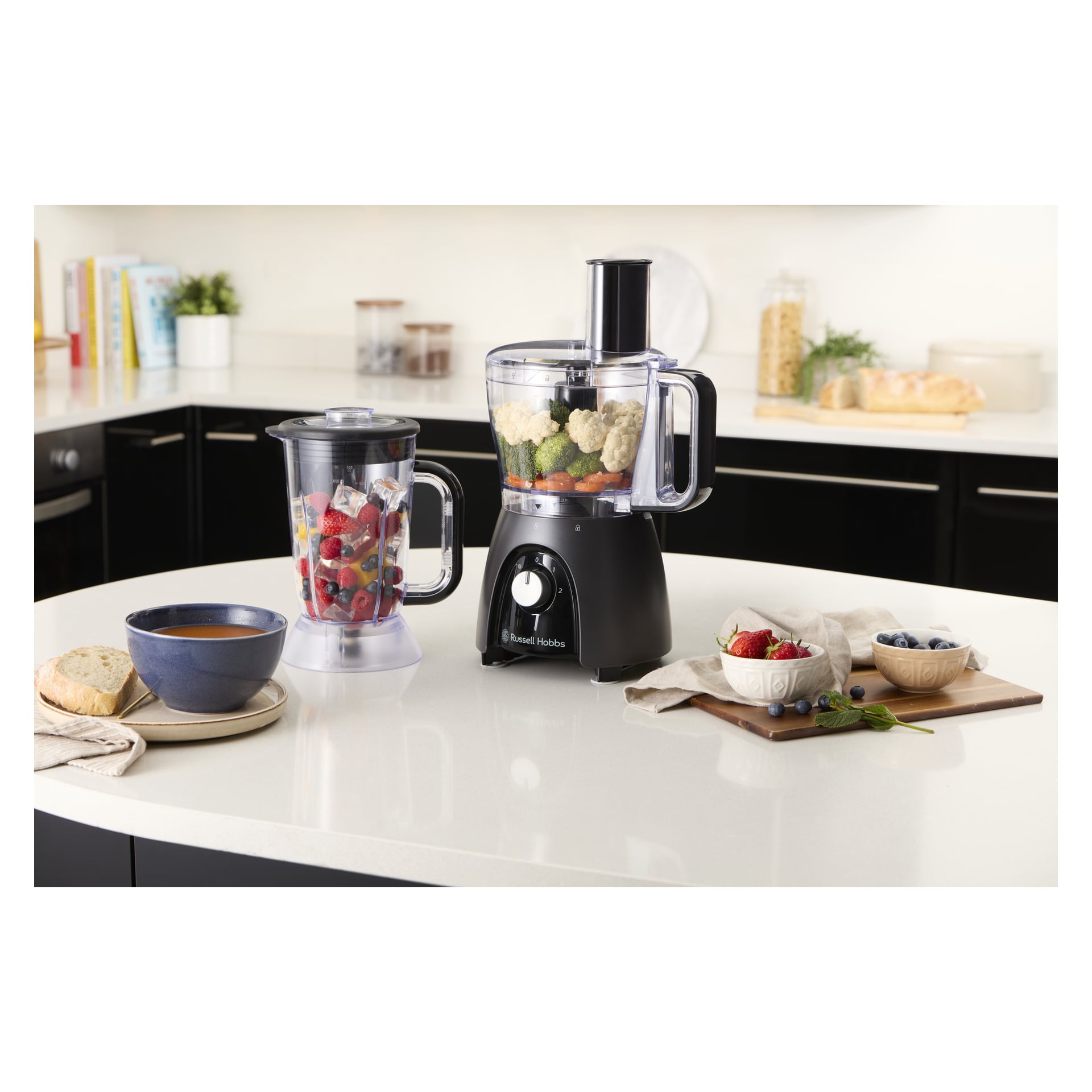 En Russell Hobbs matberedare med en mixerkanna full av frukt och grönsaker står på köksbänken, bredvid soppa, bröd och bär.