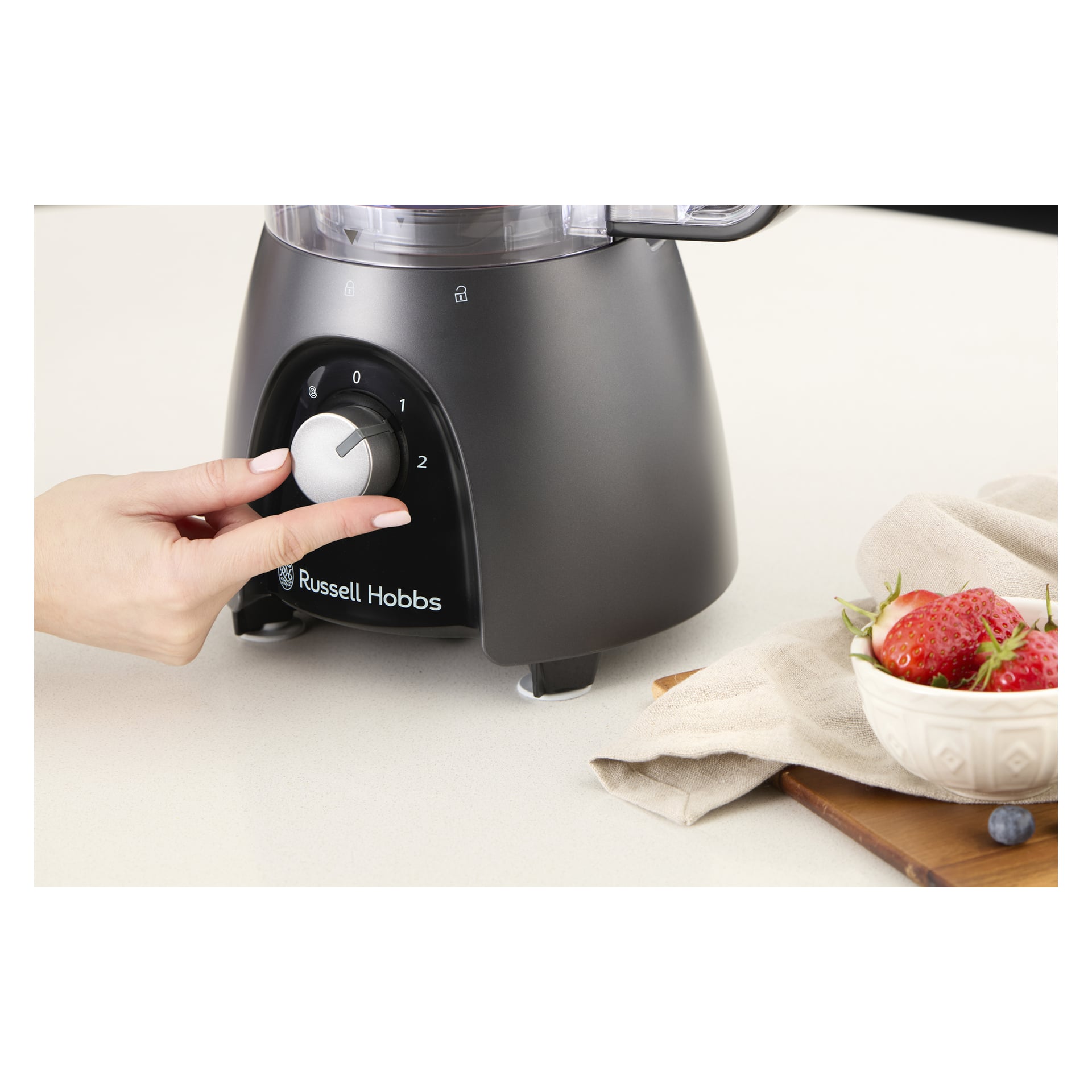 En hand justerar en Russell Hobbs matberedare med mixerkanna på en bänkskiva; I närheten finns en bricka med jordgubbar och en servett.