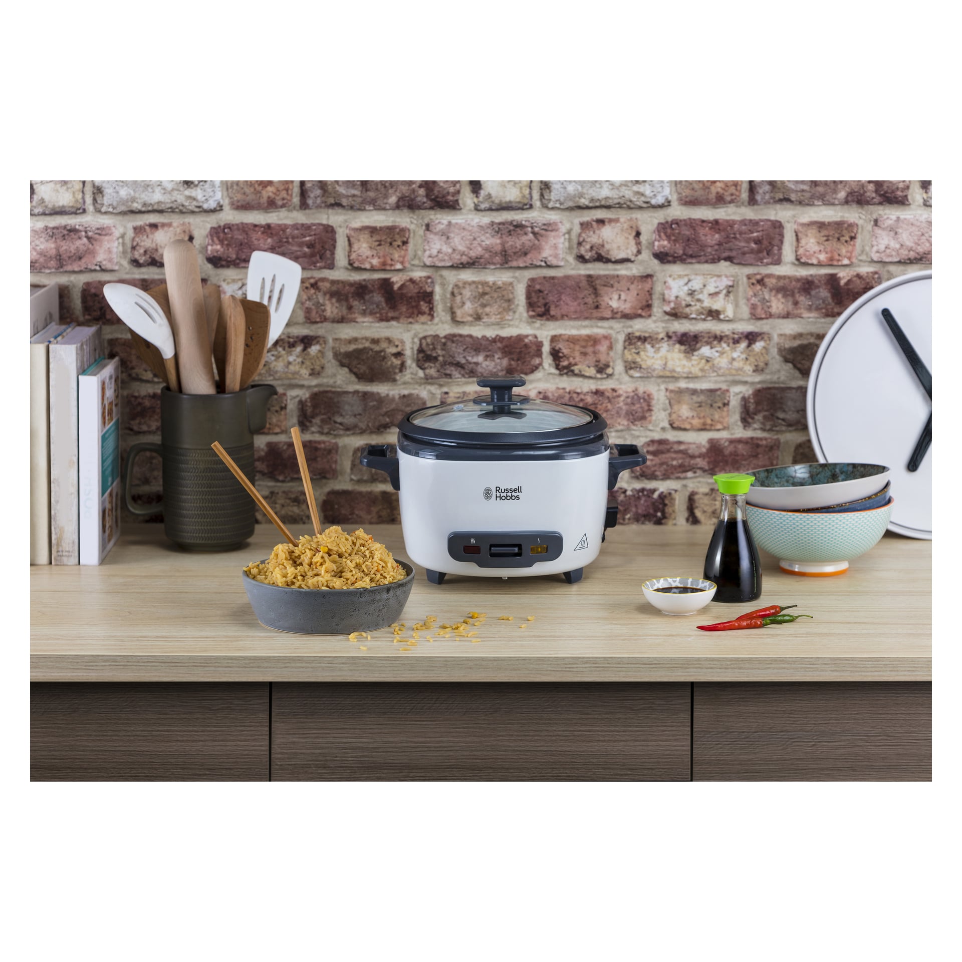 Ryżowar Russell Hobbs Duży na kuchennym blacie z makaronem, pałeczkami, przyprawami, sosem sojowym, sztućcami i książkami kucharskimi.