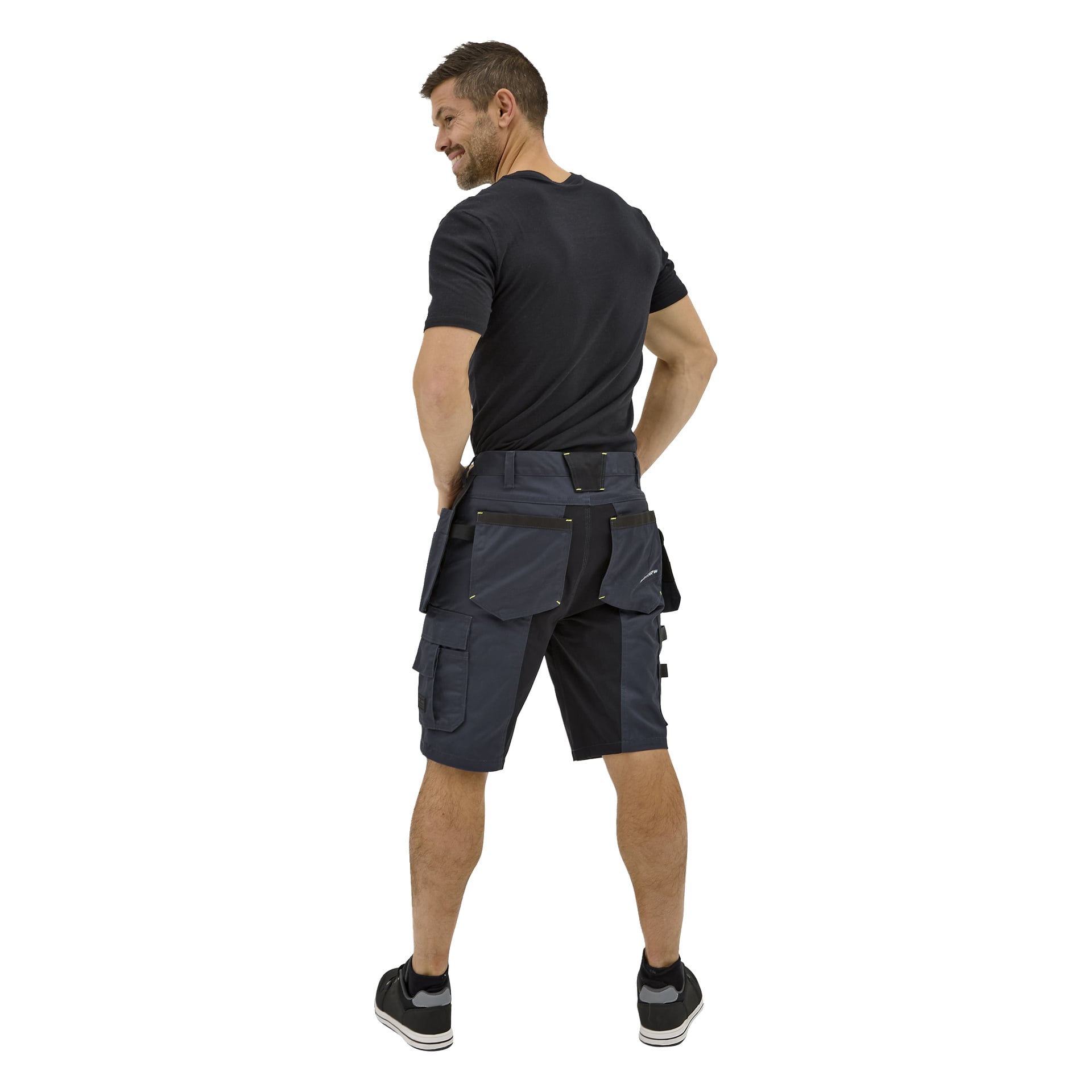 En mann sett bakfra i en svart t-skjorte, svarte joggesko med hvite såler og mørk Bluewear Rex arbeidsshorts med flere lommer.