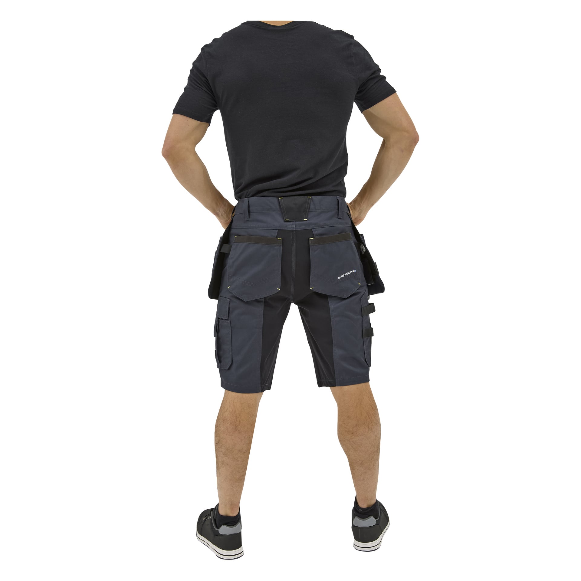 En person står med händerna på höfterna, sedd bakifrån, i Bluewear Rex arbetsshorts för herrar, en svart t-shirt och svarta sneakers.