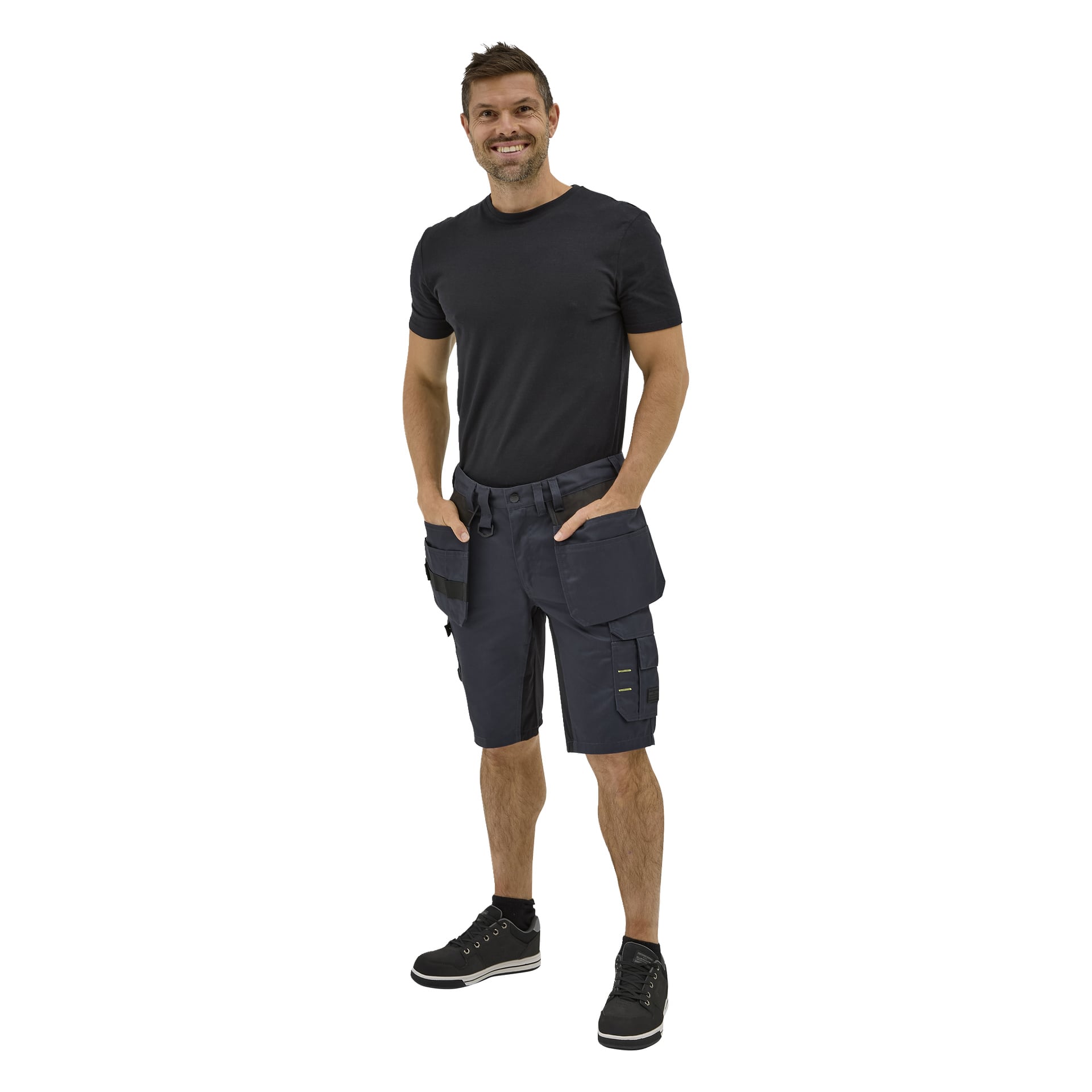 En man ler med händerna i fickorna på Bluewear Men's arbetsshorts Rex, iklädd en svart t-shirt och svarta sneakers.