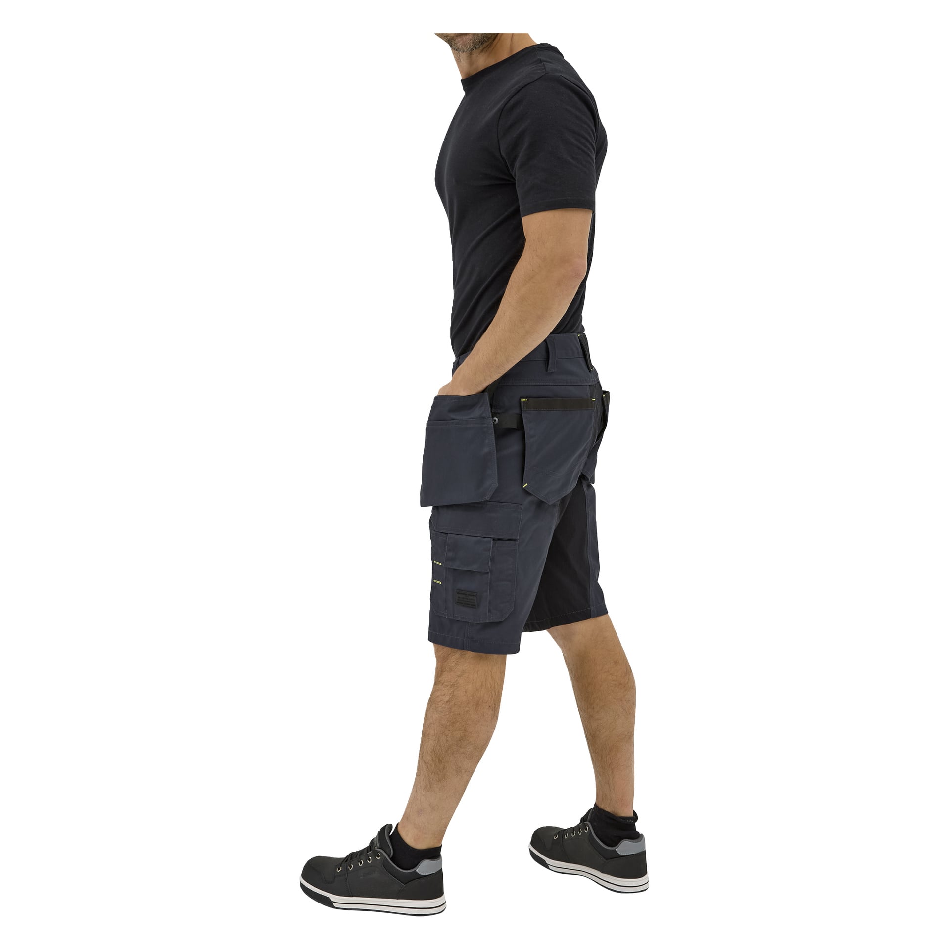 En man i profil bär en svart t-shirt, Bluewear "Men's work shorts Rex" med flera fickor och svarta sneakers. Hans vänstra hand är i fickan medan hans högra arm hänger längs sidan.