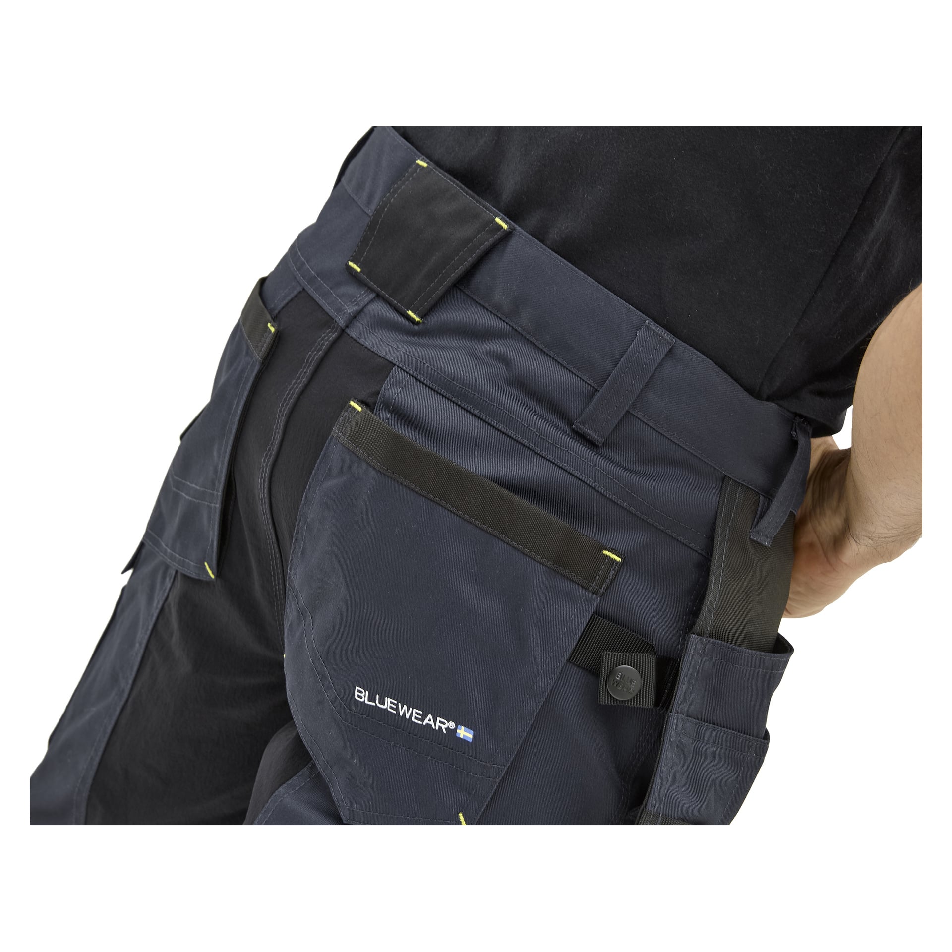 Närbild på en person i marinblå Bluewear herrshorts med svarta och gula detaljer, flera fickor och en Bluewear-logga. Svart skjorta.