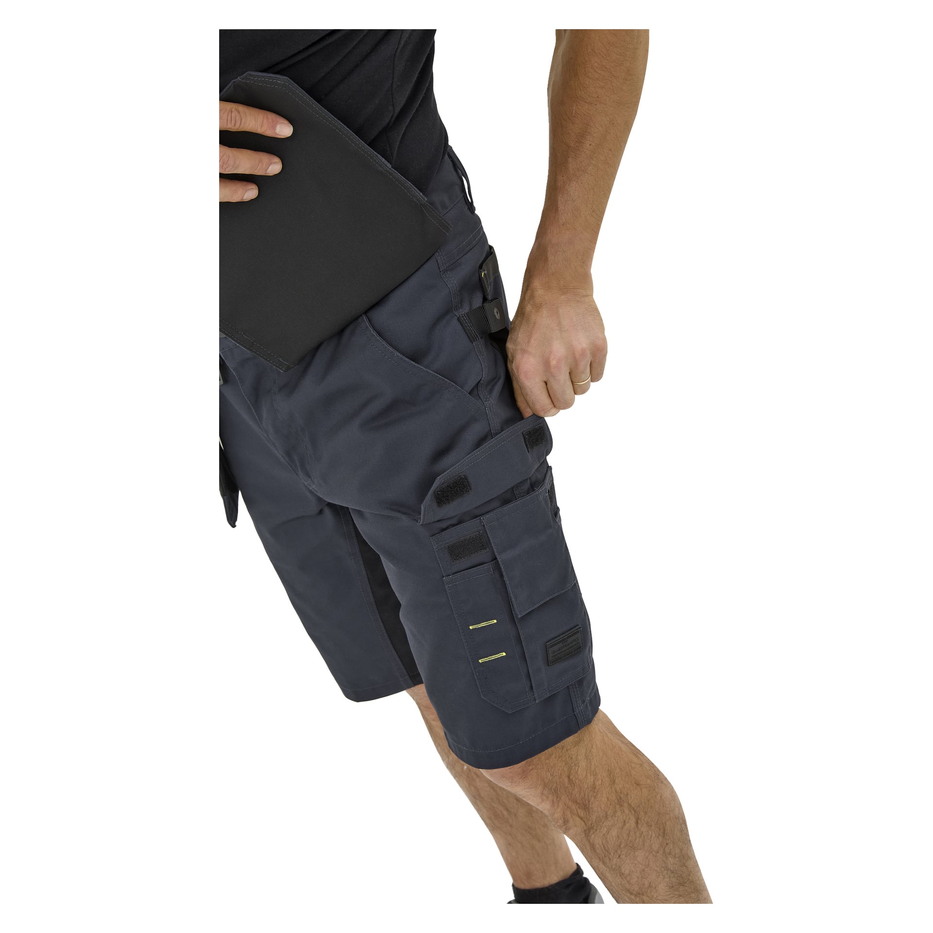 En person i Bluewear Rex arbetsshorts för herrar med flera fickor och remmar, samt en svart skjorta, håller en svart surfplatta under vänster arm.