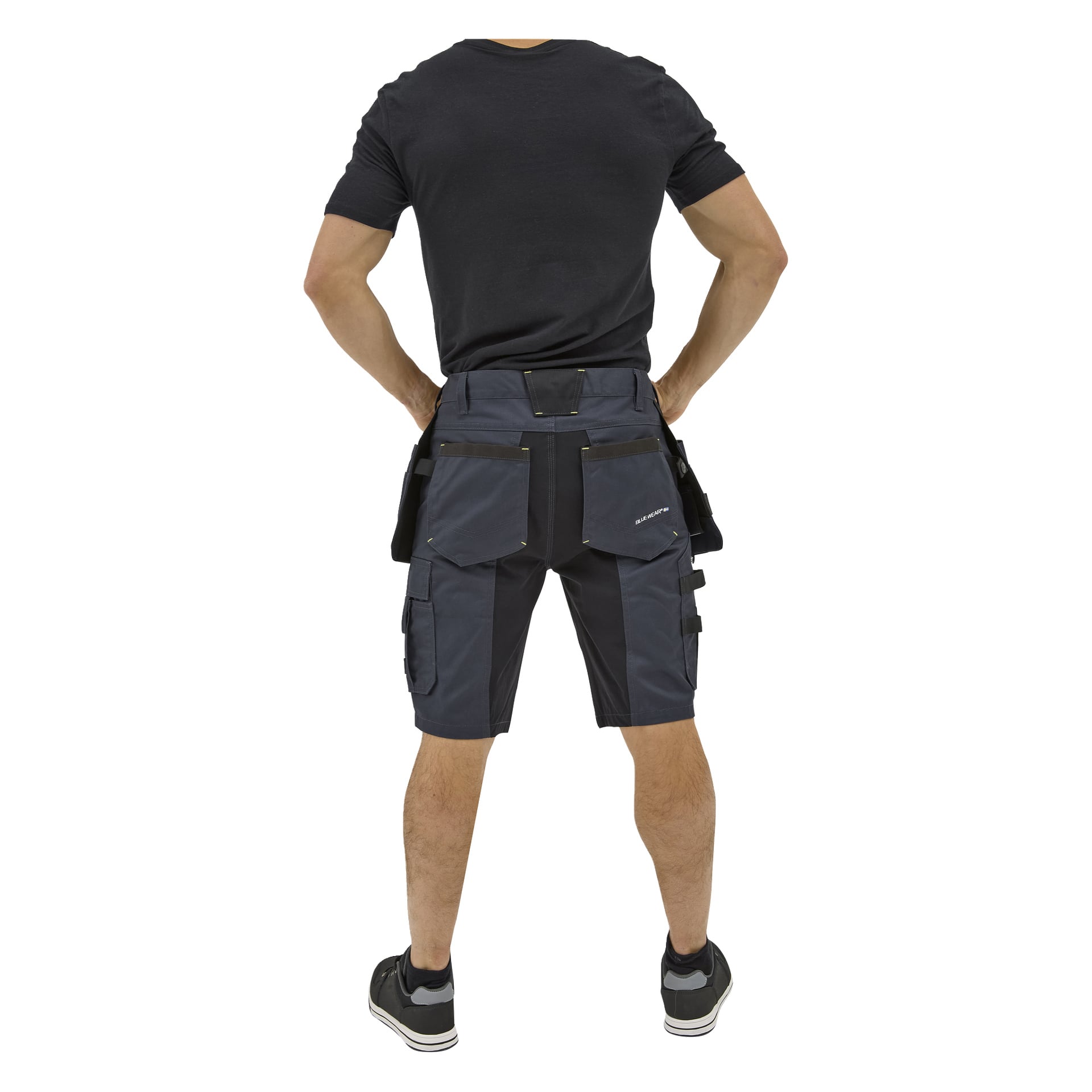 En person som ses bakifrån står med händerna på höfterna, klädd i svart t-shirt, svarta sneakers och mörka Bluewear-arbetsshorts med fickor.