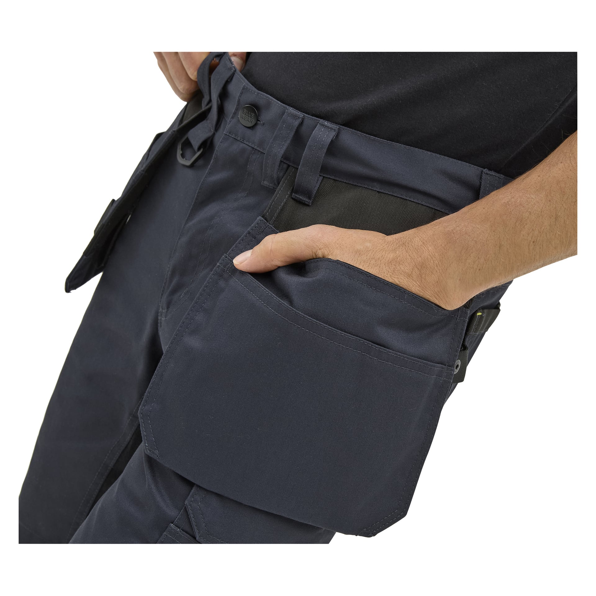 En person i Bluewear Men's Rex-arbetsshorts med svart midjeband stoppar höger hand i en stor framficka; Shorts har många fickor.