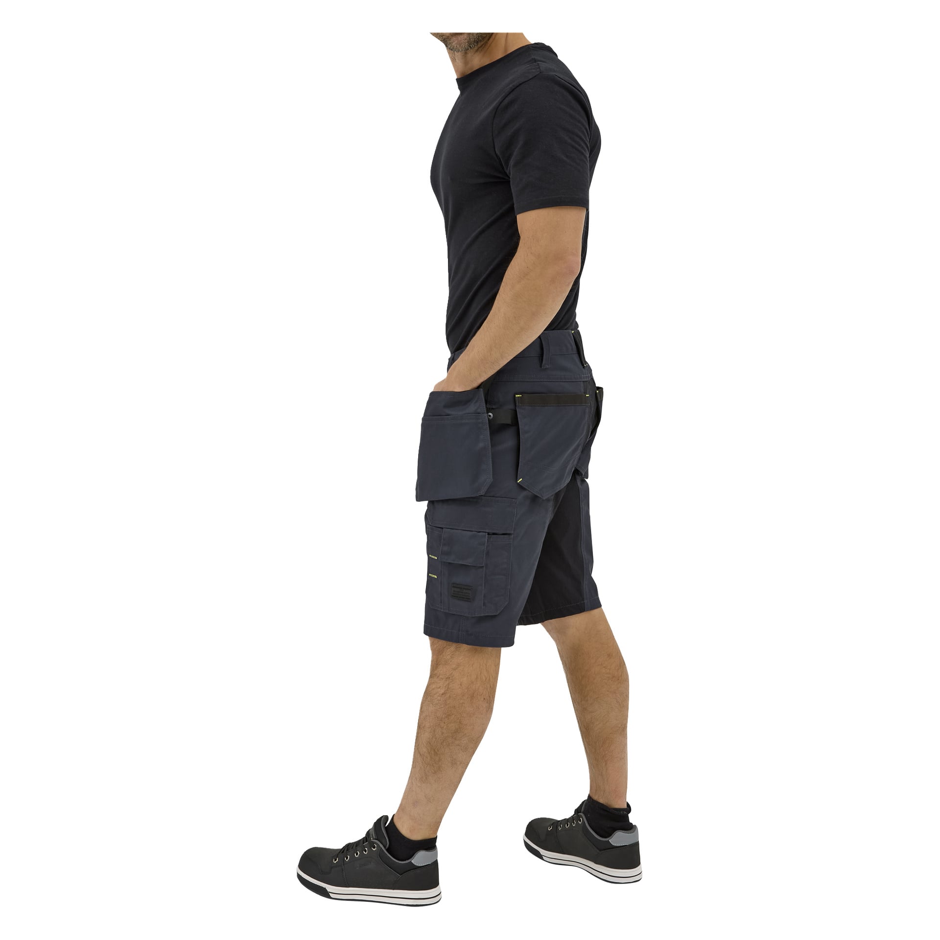 En person går i en svart t-shirt, Bluewear Men's Rex arbetsshorts med många fickor och verktygsfickor, samt svarta sportskor, sedda från sidan.