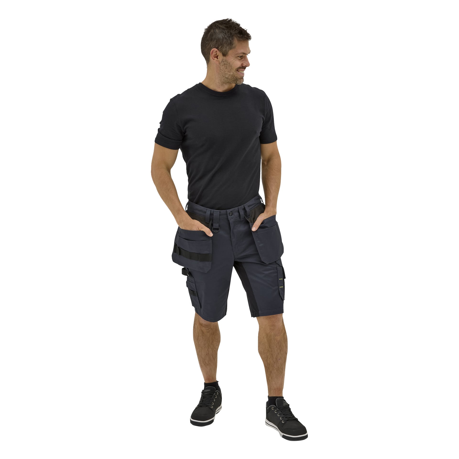 En man ler åt sidan med händerna i fickorna, klädd i mörkblå Rex-arbetsshorts, en enkel svart t-shirt och svarta sneakers.