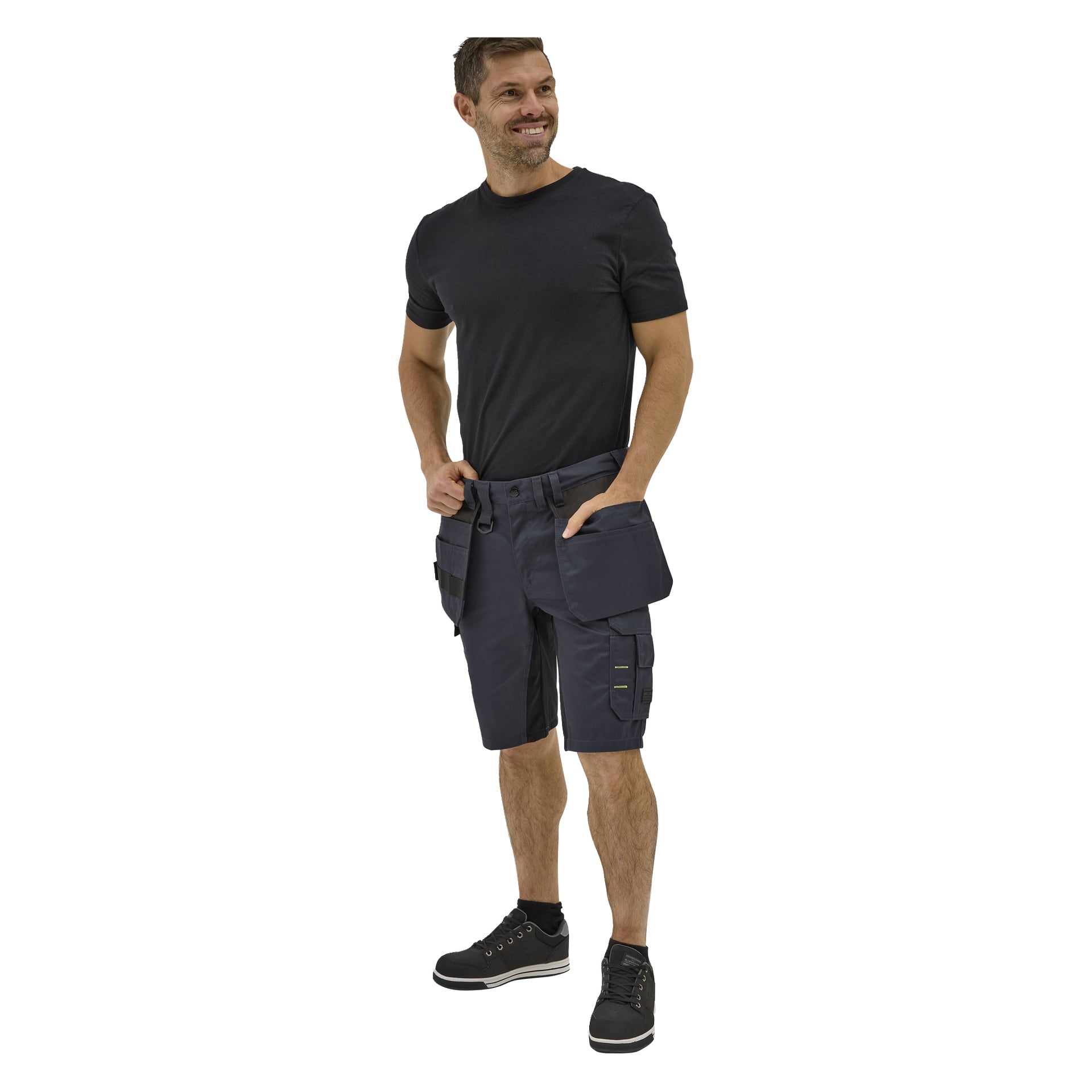Leende man i svart t-shirt, Bluewear-arbetsshorts med fickor och öglor, och svarta sneakers; en hand i fickan.