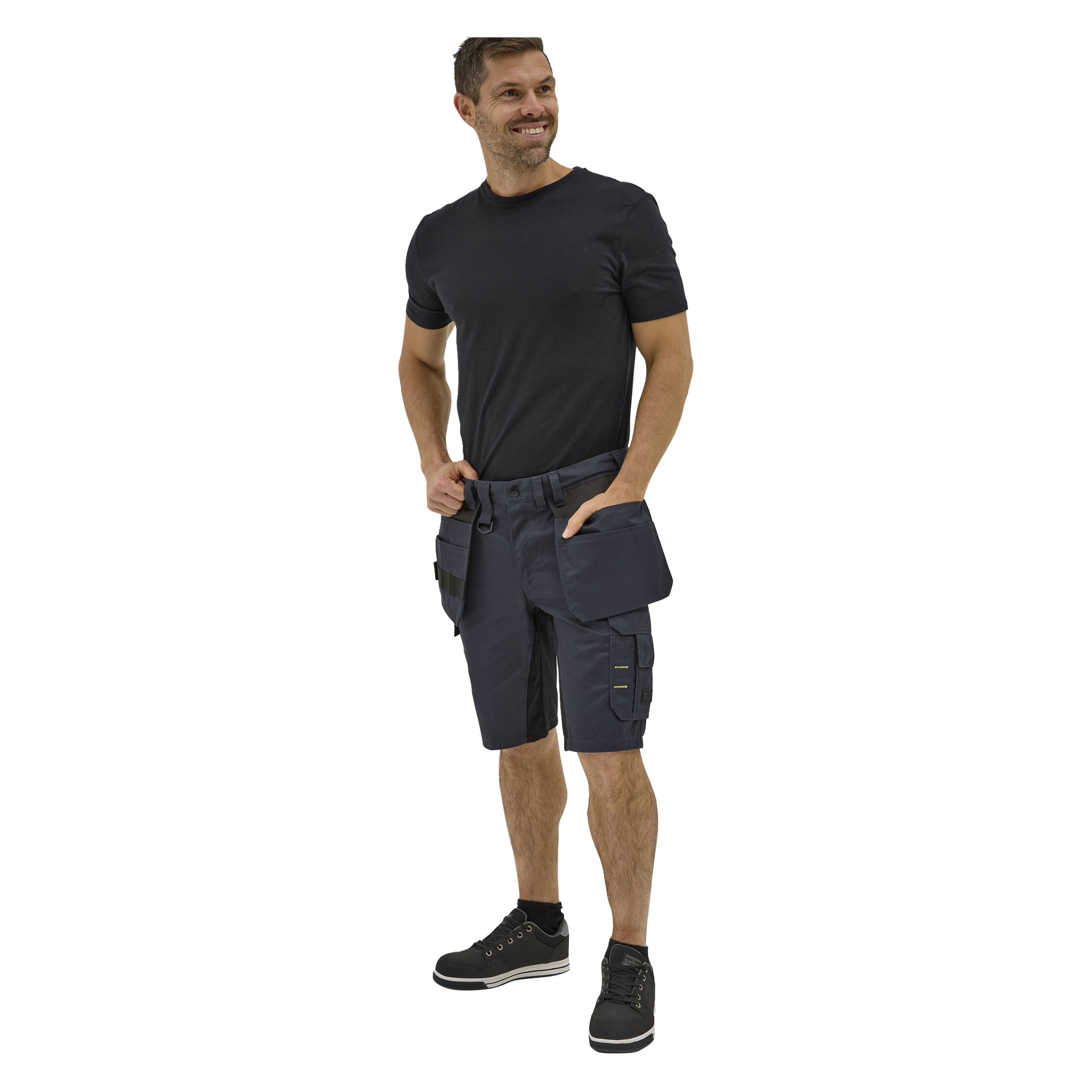 En leende man poserar med händerna i fickorna på Bluewear Rex-arbetsshorts, iklädd en svart T-shirt och svarta sneakers med vita sulor.