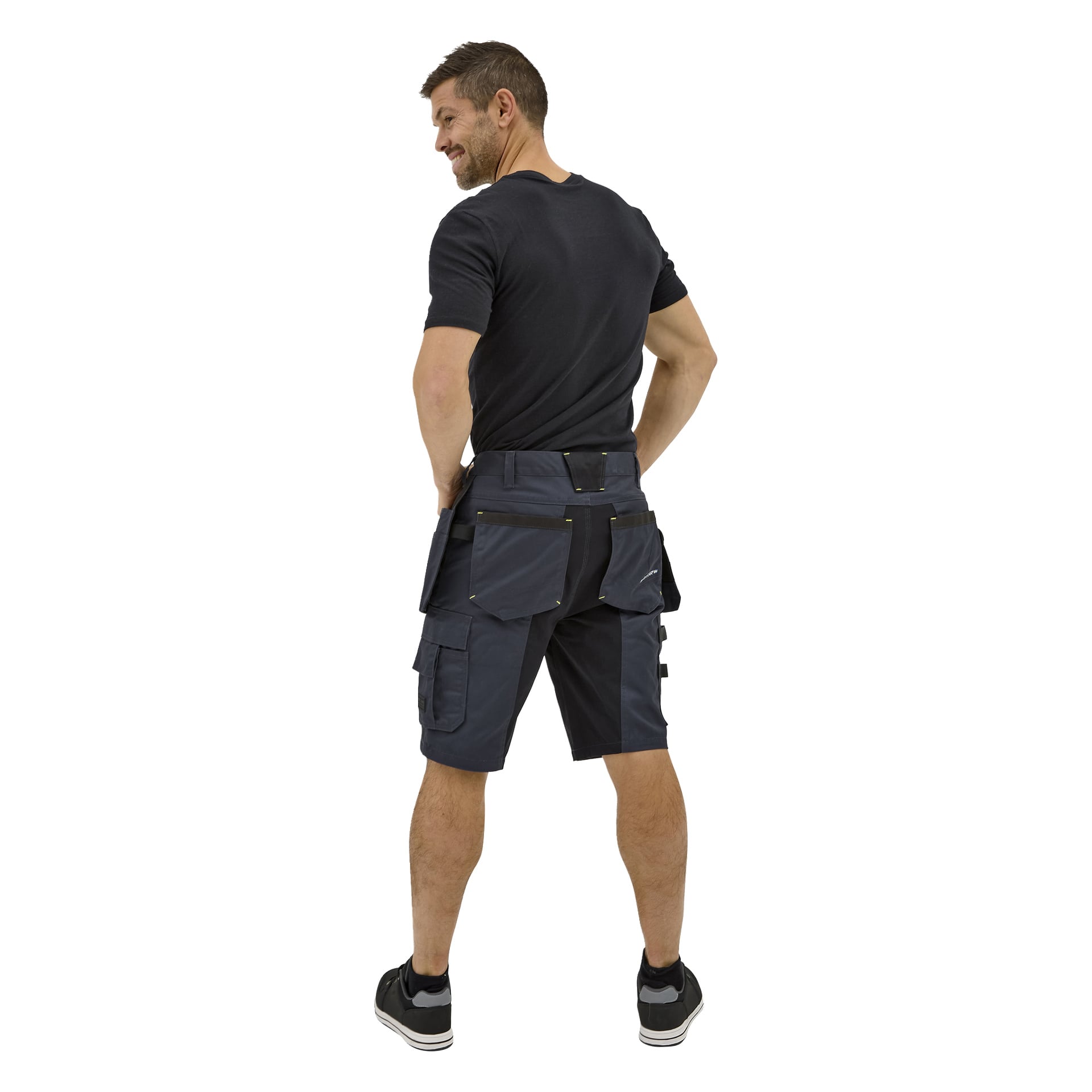 En leende man bär Bluewears mörka Rex-arbetsshorts med flera fickor tillsammans med en svart t-shirt och svarta sneakers.