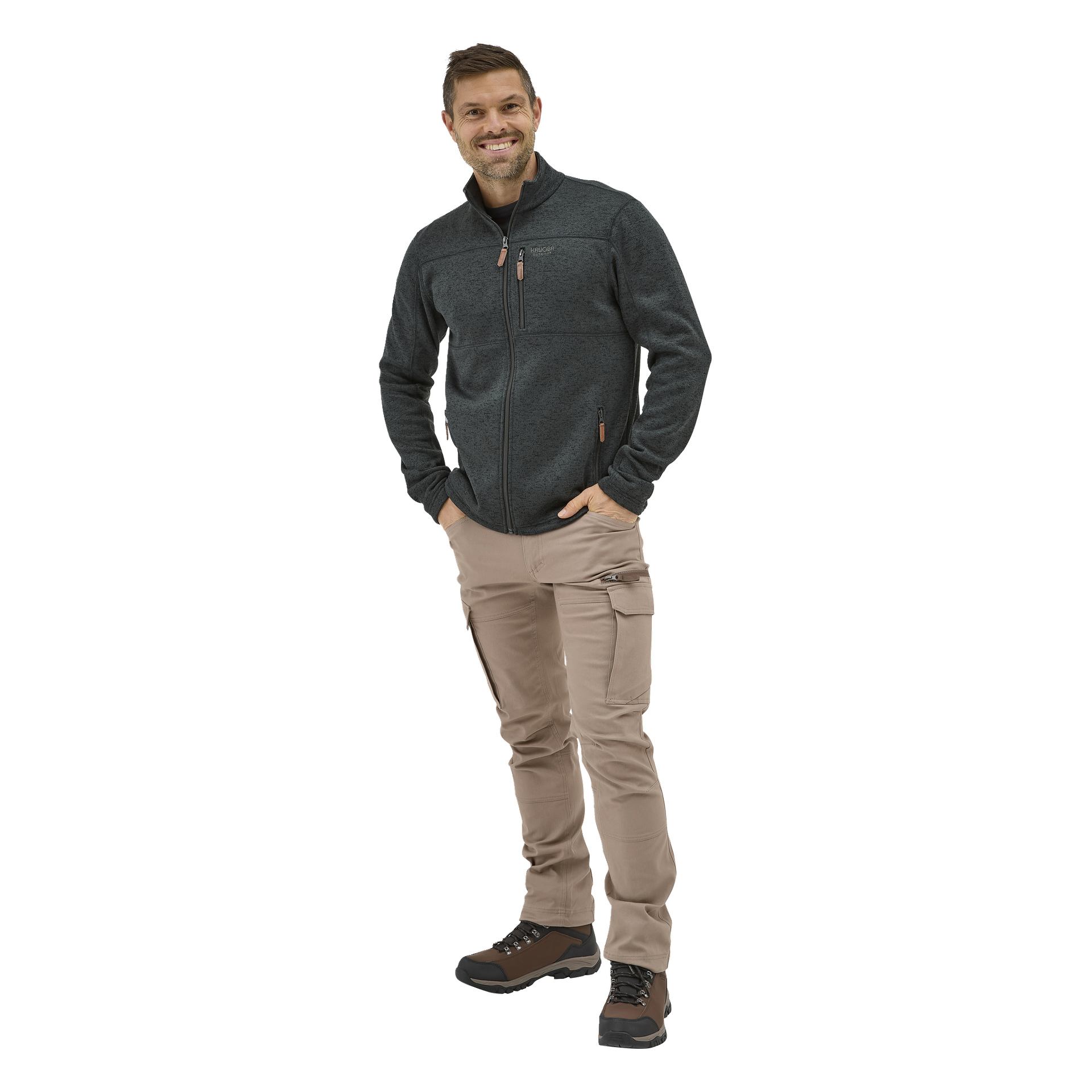 Ein lächelnder Mann mit den Händen in den Taschen trägt Kayoba Outdoors Åsen-Hose, eine dunkle Reißverschlussjacke und braune Wanderschuhe.