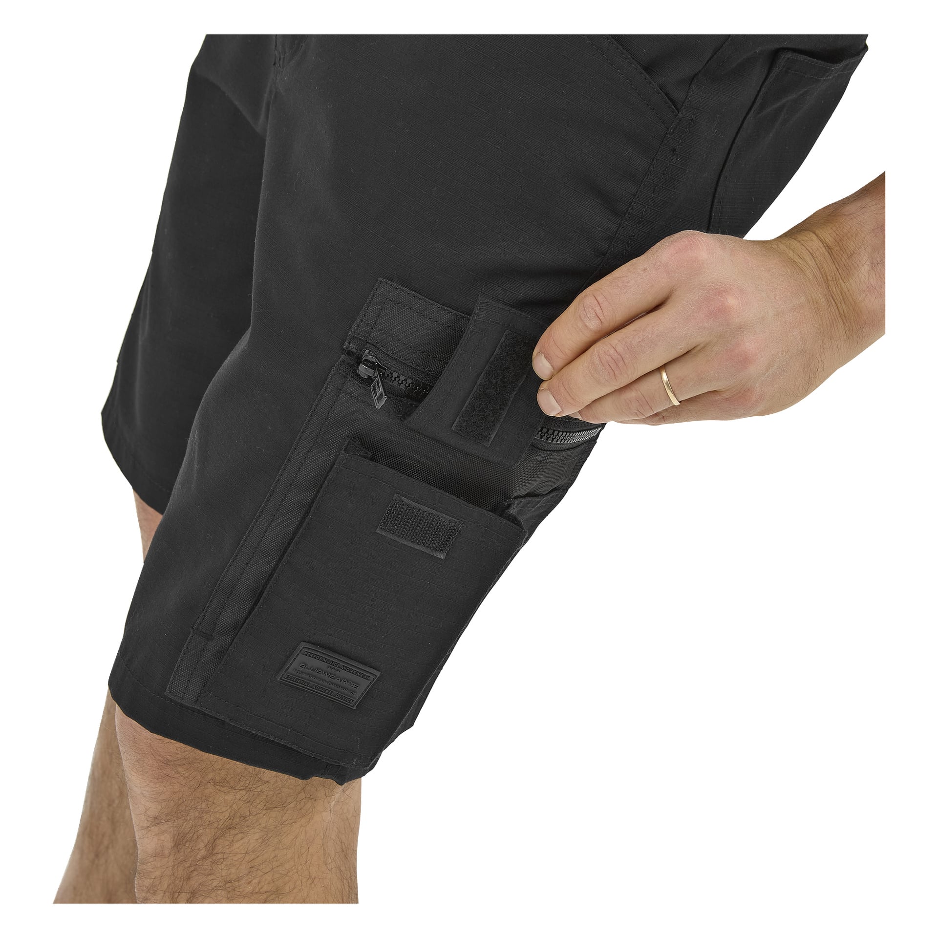 En person i Bluewear Micke arbetsshorts öppnar en sidoficka med kardborreband, som visar en ficka med dragkedja ovanför och bär en guldring.
