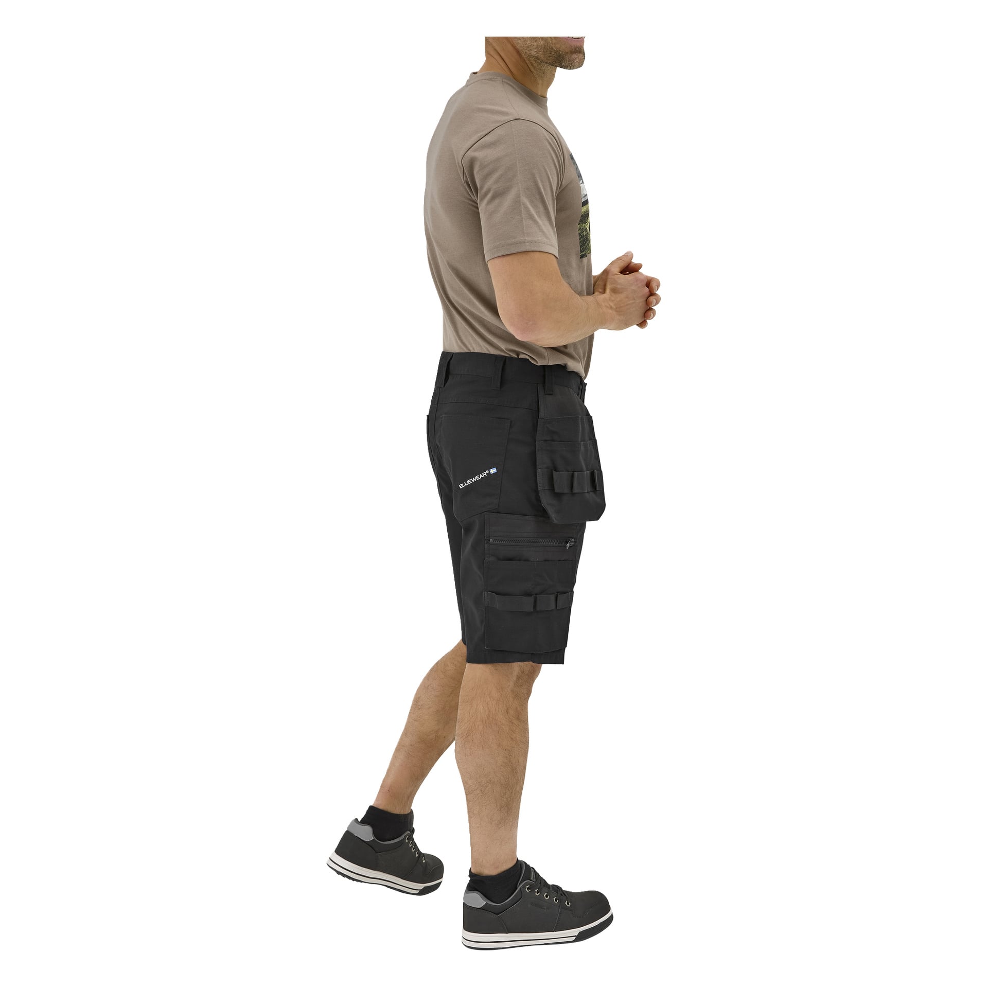 En man i sidoprofil bär en taupe t-shirt, svarta cargoshorts för arbete, svarta strumpor och svarta sneakers. Händerna knäppta framför sig.