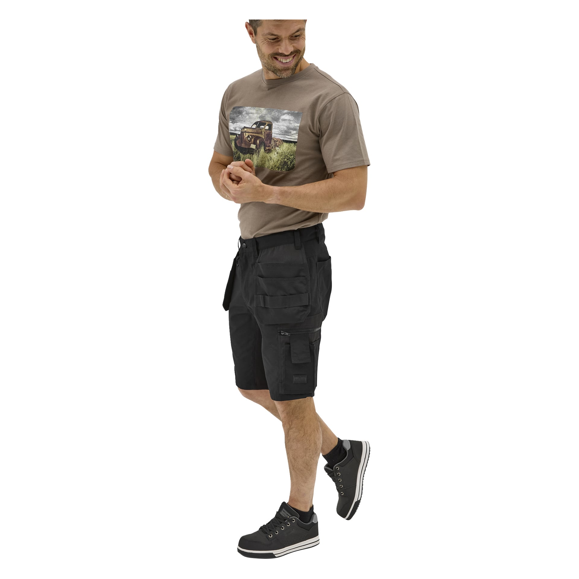 En leende man bär en taupe grafisk t-shirt med en lastbil, svarta Bluewear Micke arbetsshorts med fickor och svarta sneakers med vita sulor.