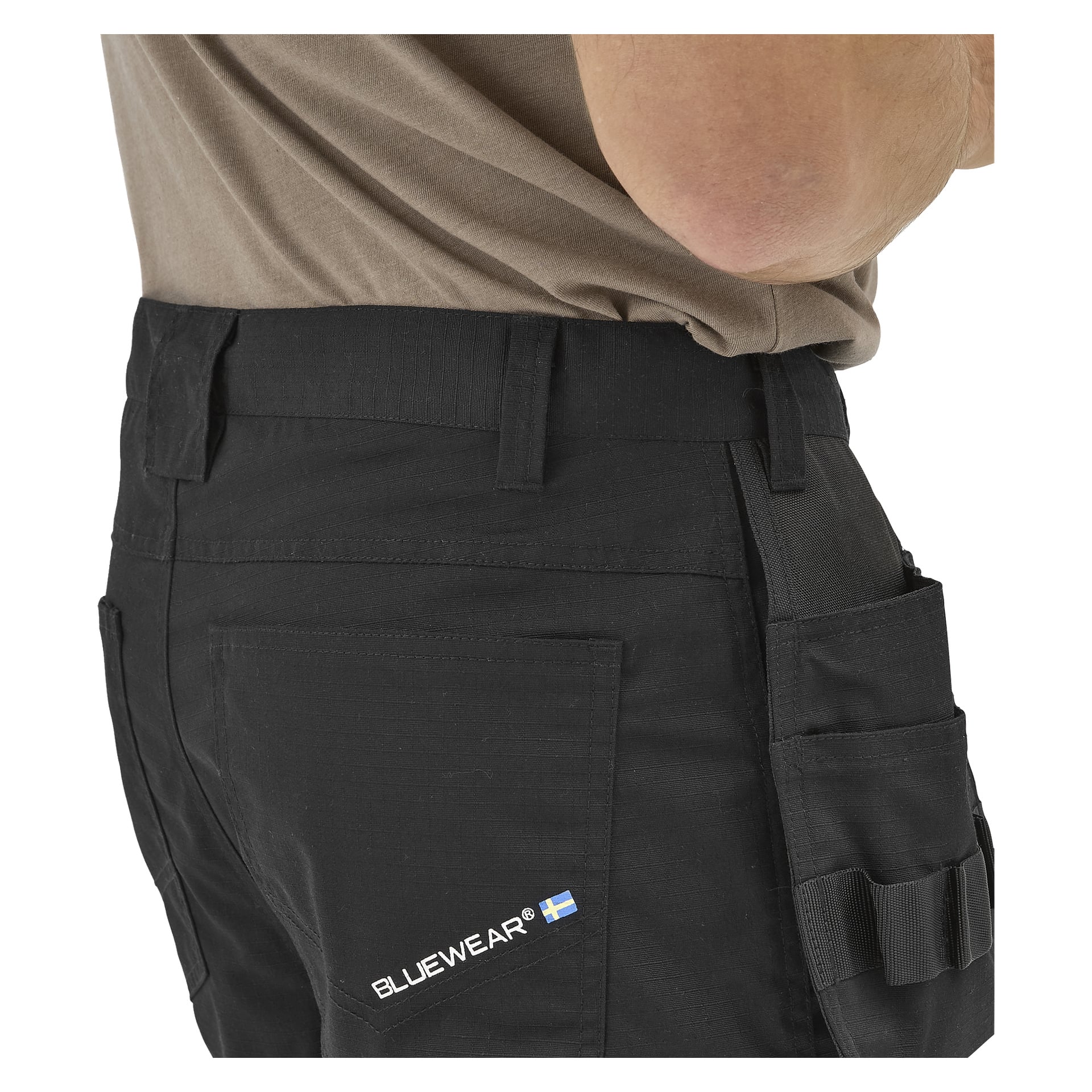 En närbild från sidan av en person i Bluewear herrshorts från Micke arbete, logga och liten svensk flagga på bakfickan, beige skjorta.