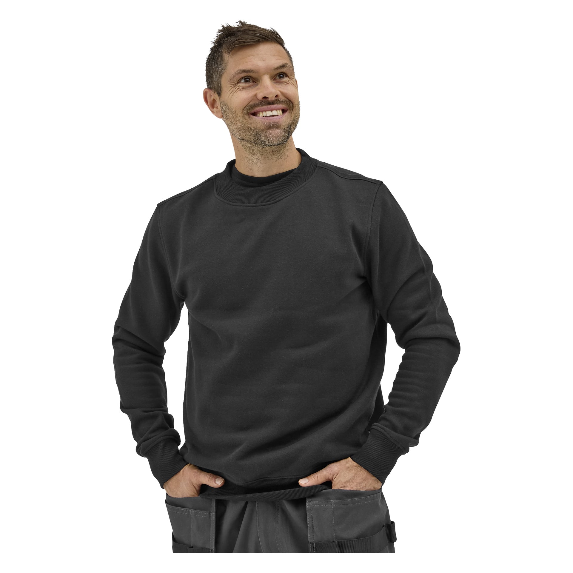 En leende man med kort brunt hår och skägg bär Bluewear Men's Sweater Noa och grå byxor, vänd lite åt vänster, händerna i fickorna.