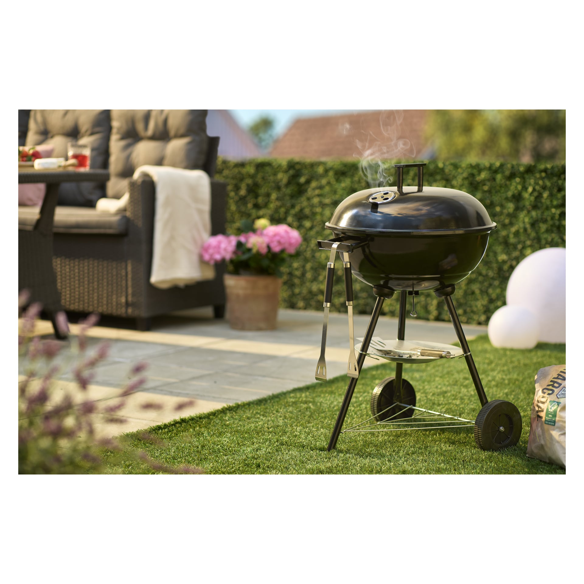 Burns & Barkles Kettle grill Coal King 430 na trawiastym patio, sztućce wiszą, unoszący się dym, sofa, doniczka i żywopłoty z tyłu.