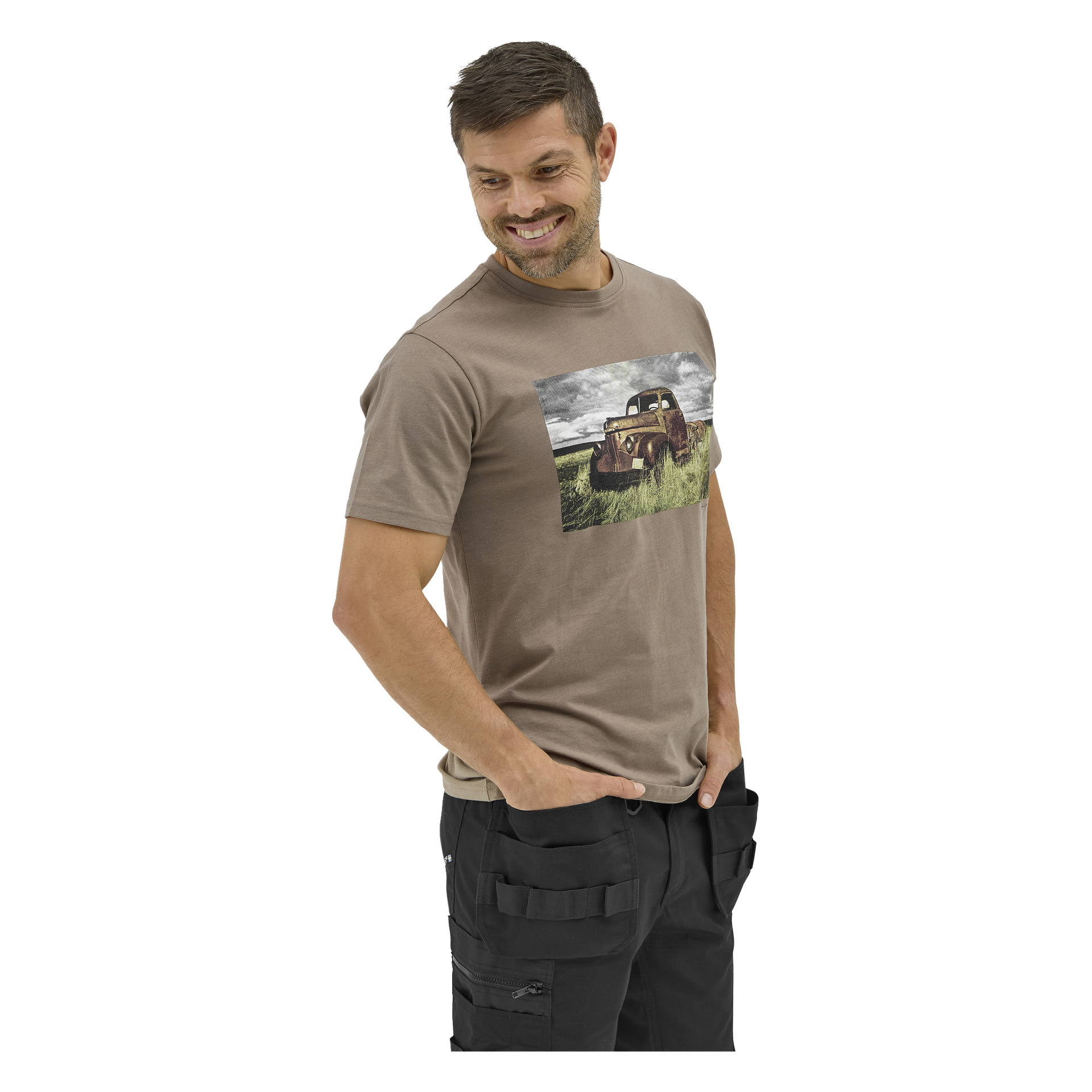 En skjeggete mann med kort hår smiler, ser ned, iført en Bluewear Sebbe T-skjorte med en grafikk av en rusten lastebil i gresset.