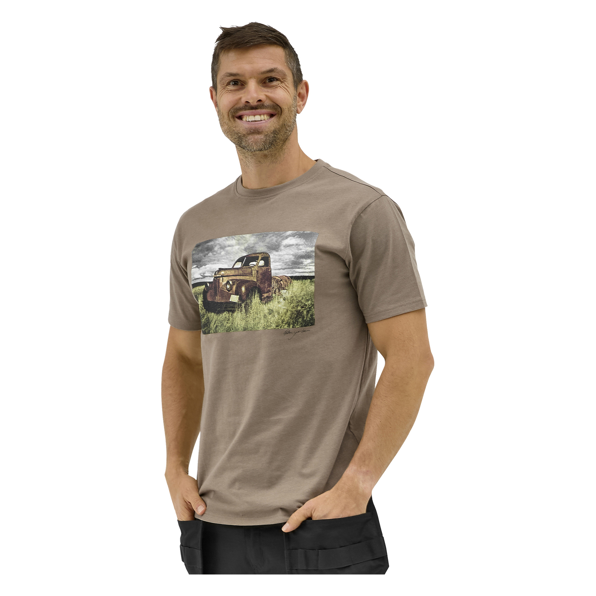 En leende man med kort brunt hår och skägg bär en Bluewear Sebbe-T-shirt som visar en gammal lastbil på en gräsbevuxen äng och med molnig himmel.