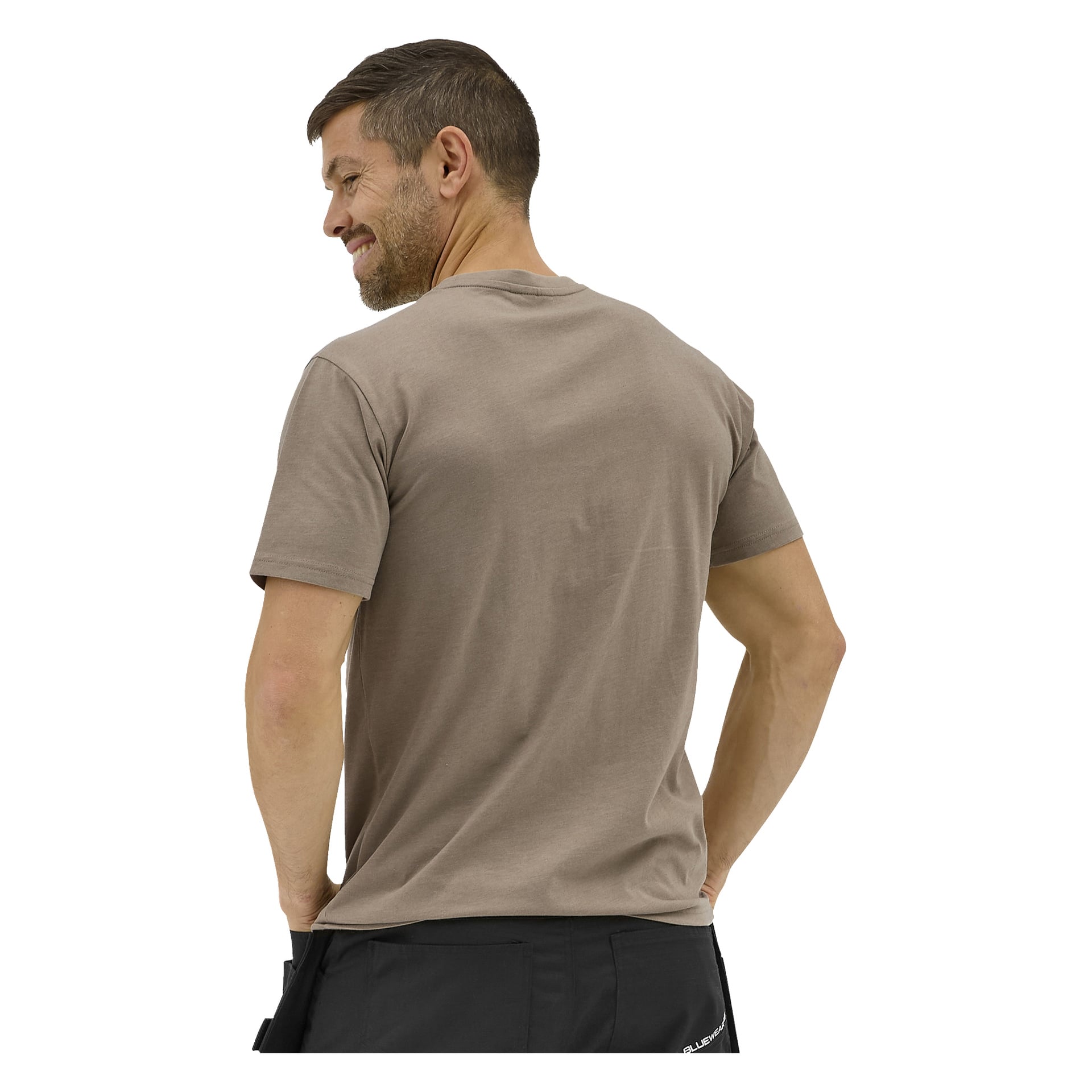 En man med kort brunt hår och skägg vänder bort blicken, händerna på höfterna, iklädd en taupe Bluewear Sebbe T-shirt och svarta byxor.