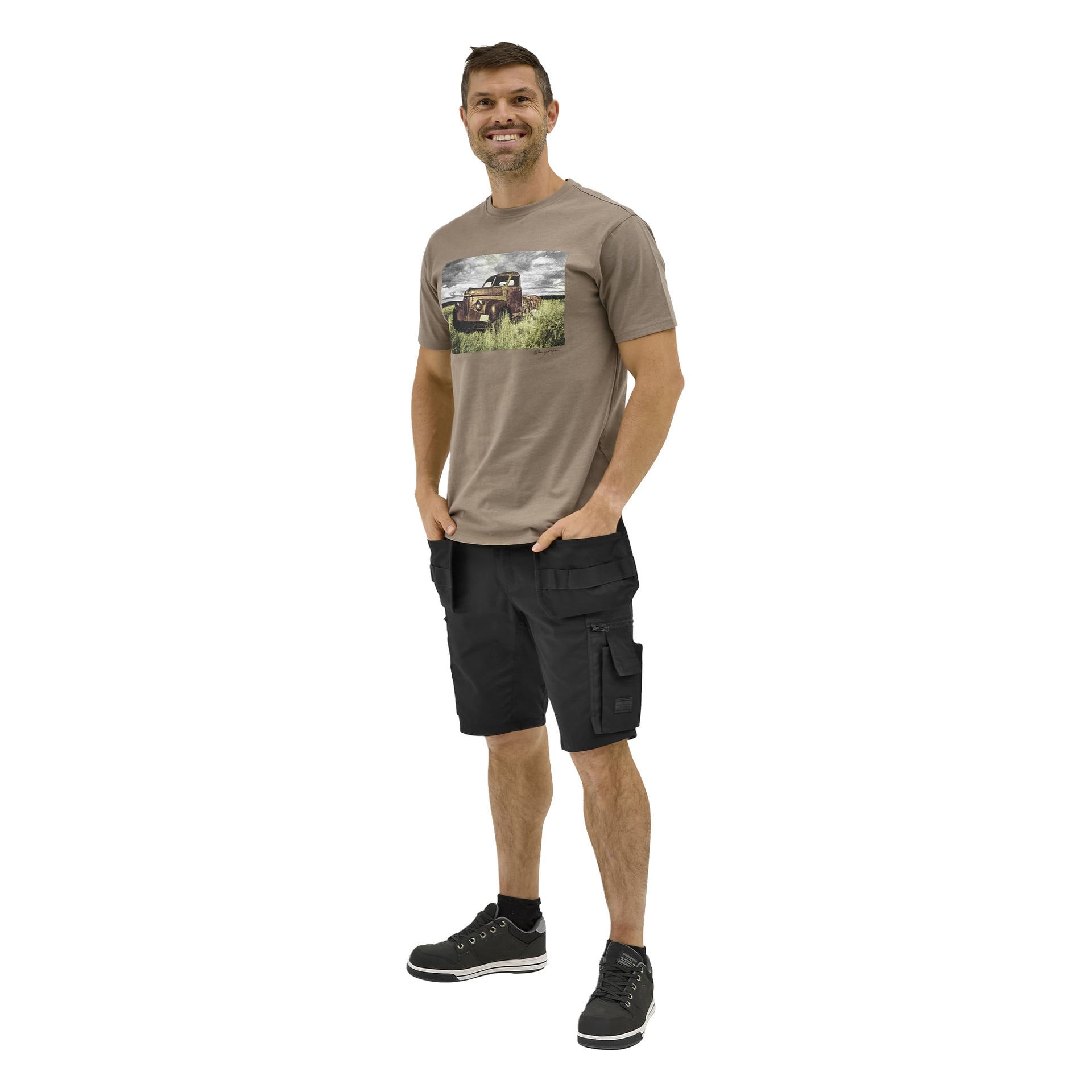 En man ler med händerna i fickorna, bär en brun lastbilströja, svarta cargoshorts och svarta sneakers.