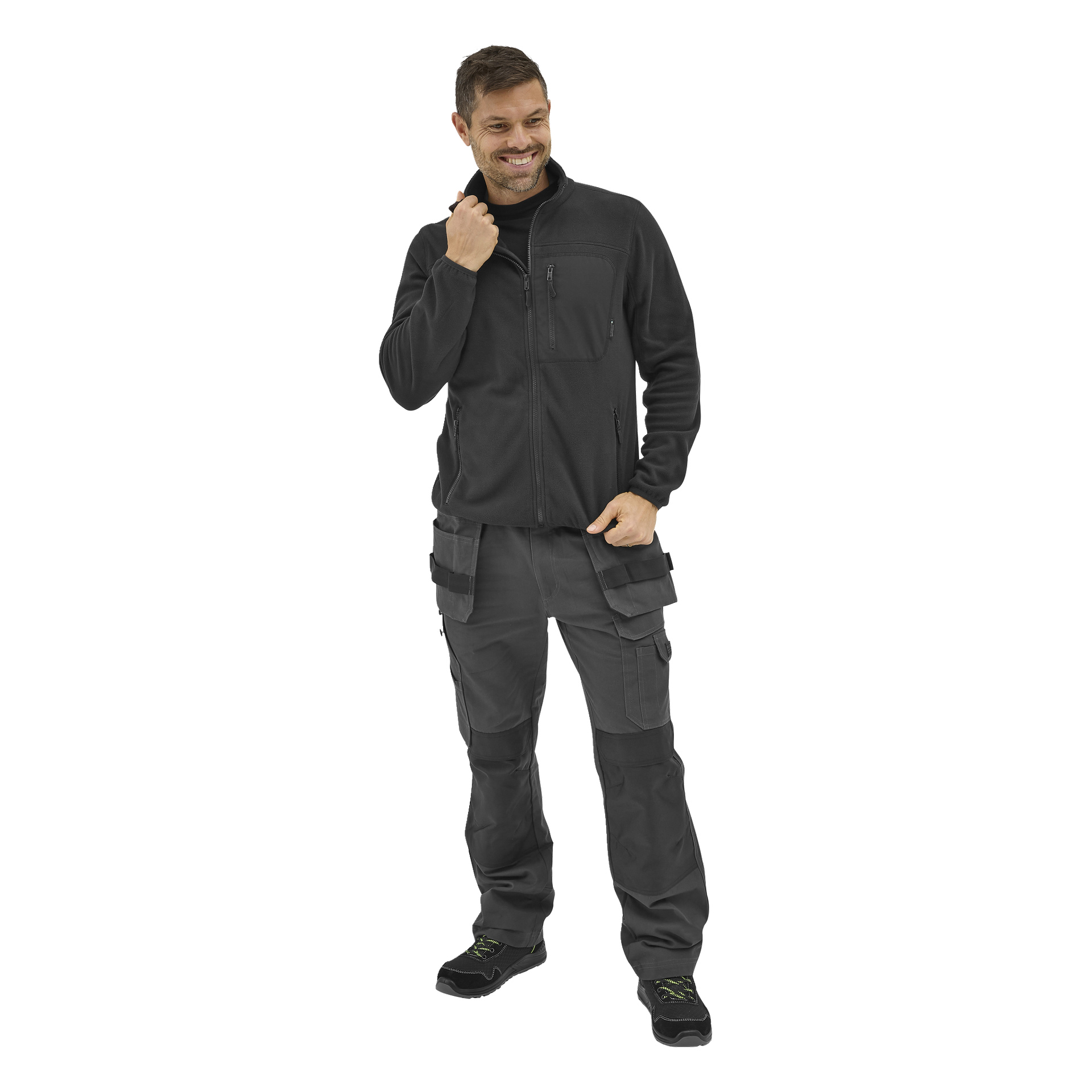 En man ler i Bluewear Men's fleece-tröja Filip med svarta cargo-arbetsbyxor och svarta arbetsskor med gröna detaljer. Han håller i tröjans dragkedja med ena handen i fickan.