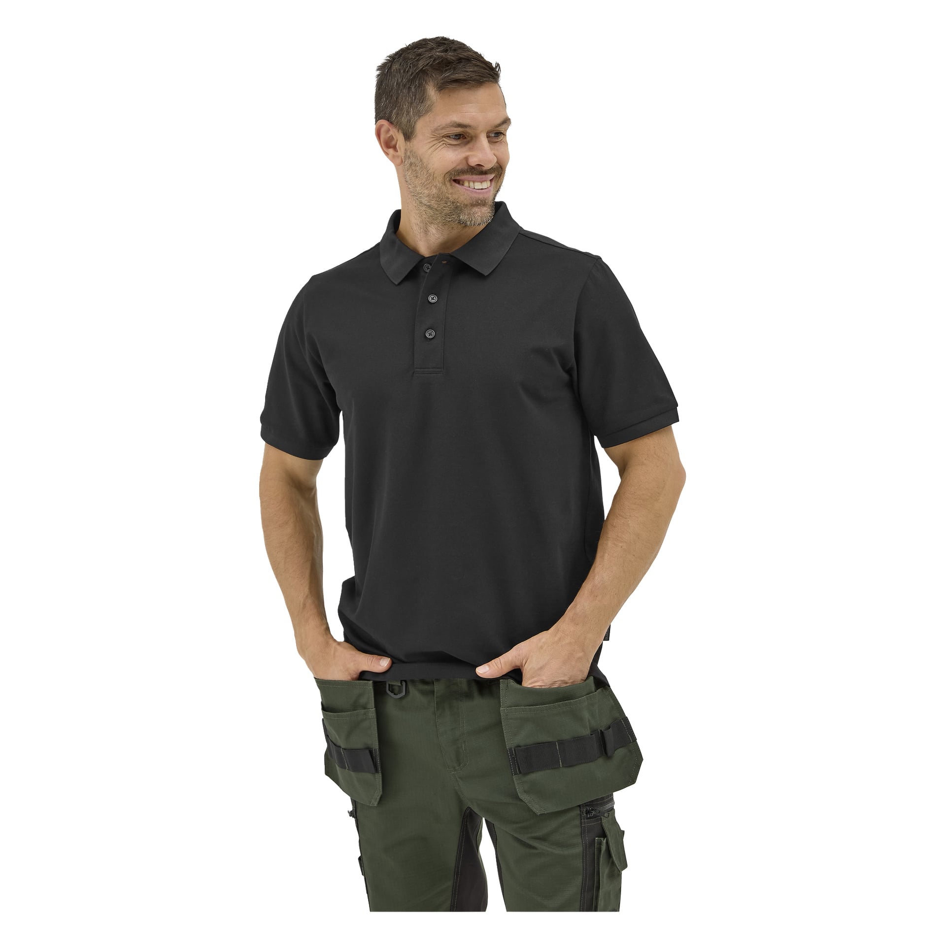 En man i svart Bluewear-pikétröja och gröna verktygsbyxor med svarta fickor står händerna i fickorna, ler och tittar åt sidan.