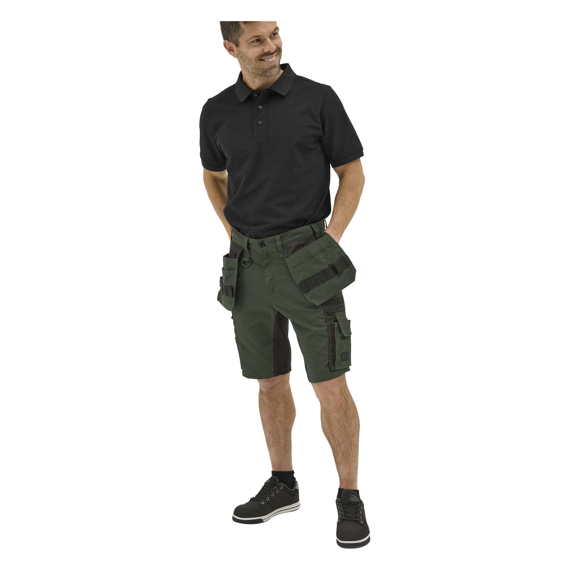 En man ler med händerna i fickorna, klädd i en Bluewear Putte herrpikétröja, gröna verktygscargoshorts och svarta sneakers.