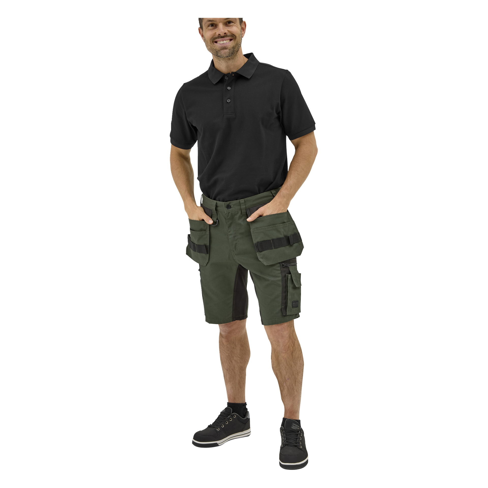 En man ler med ena handen i fickan, klädd i en Black Bluewear Putte-pikétröja, gröna arbetsshorts med flera fickor och svarta sneakers.