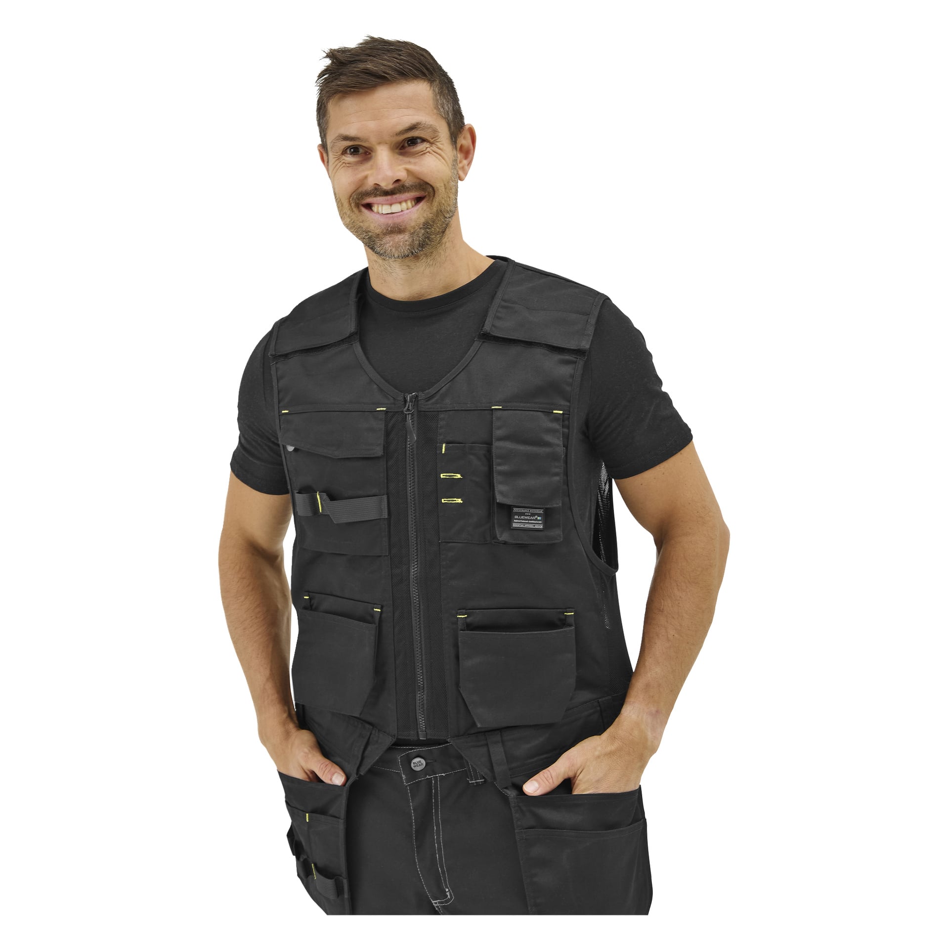 En leende man har händerna i de stora framfickorna på en Bluewear Men's arbetsväst över en svart kortärmad skjorta.
