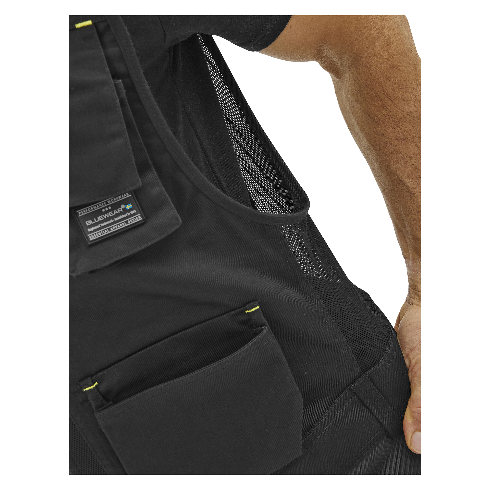 Närbild på en person i en svart Bluewear herrväst med nätpanel, flera fickor och synlig Bluewear-etikett; Höger arm delvis synlig.