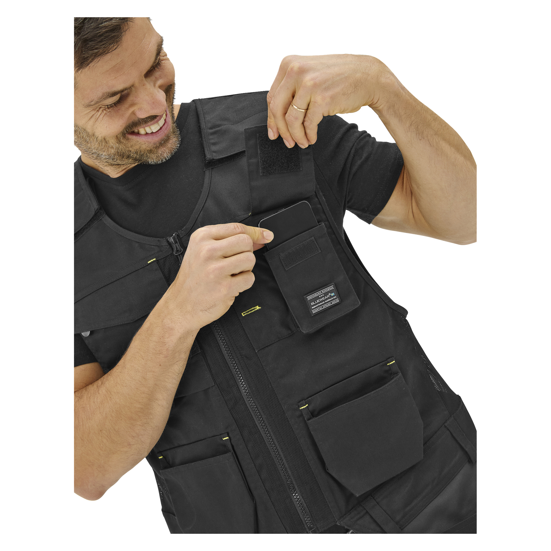 En man i en Bluewear Men's arbetsväst med flera fickor ler när han öppnar en flik och tittar ner på fickan.