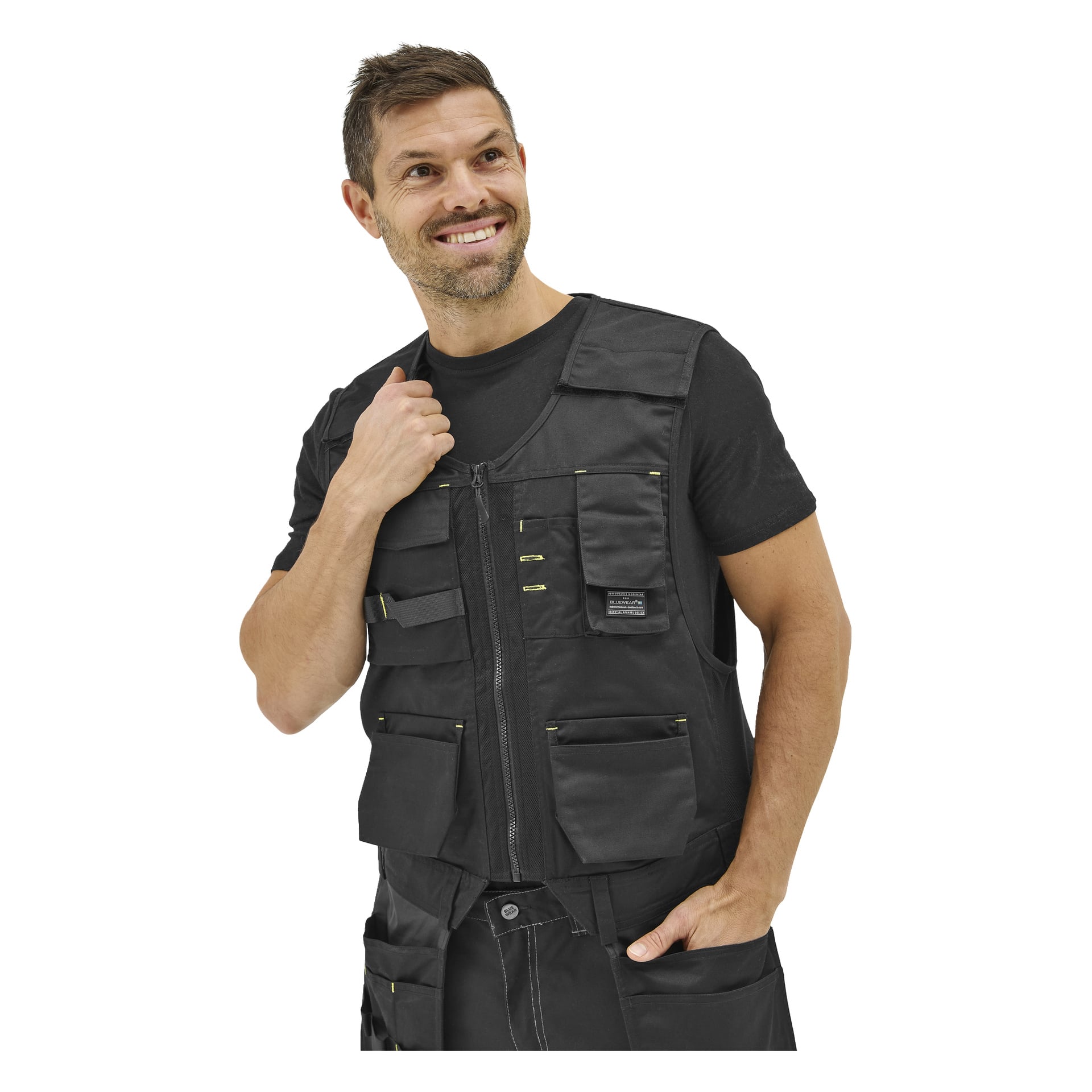 Ein Mann in einer schwarzen Bluewear-Utility-Weste mit mehreren Taschen über einem schwarzen T-Shirt steht lächelnd da, eine Hand am Kragen, und blickt zur Seite.