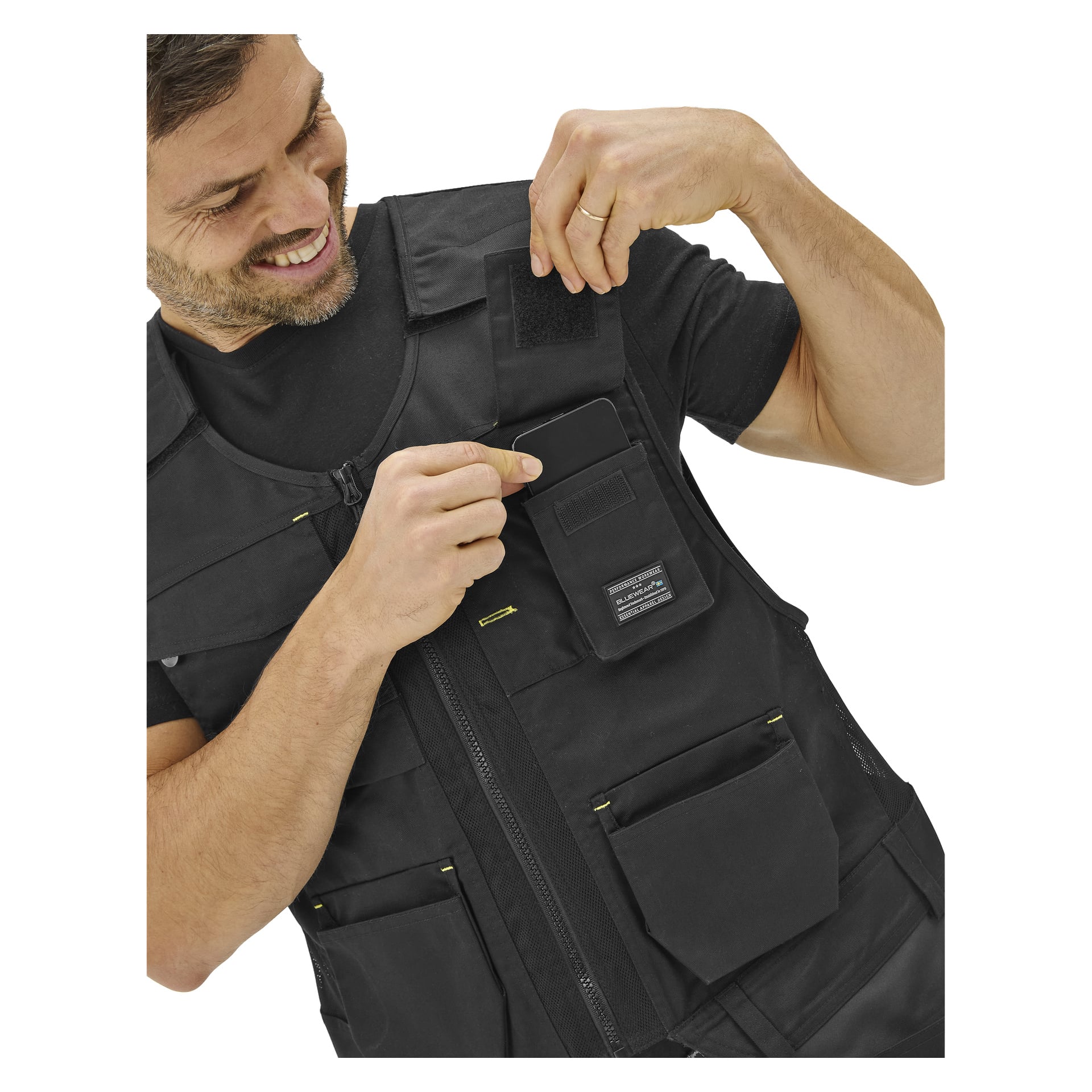 En leende man öppnar kardborrebandsluckan på en övre ficka på en Bluewear Men's arbetsväst och visar ett litet rektangulärt föremål inuti.