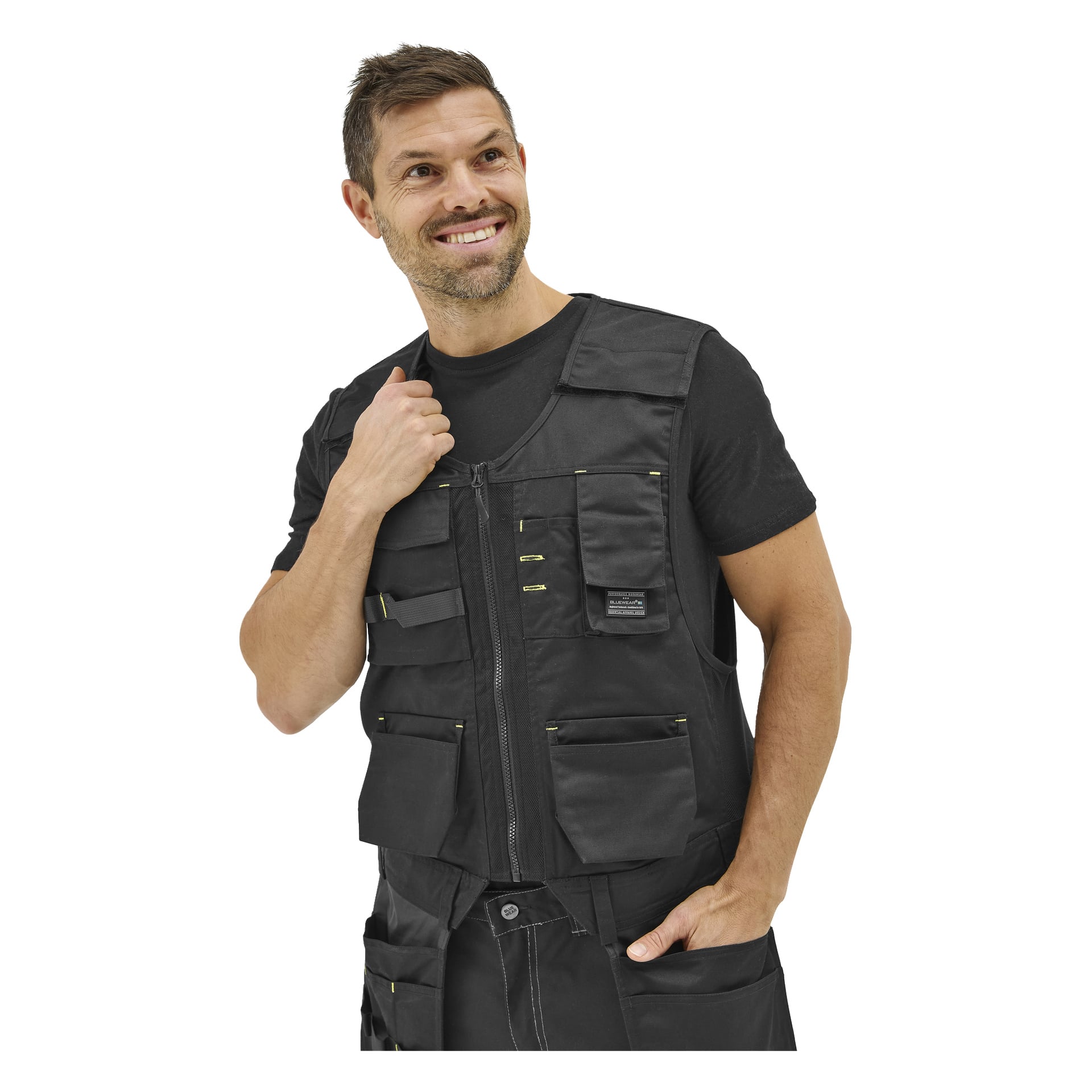 En man bär Bluewear Men's arbetsväst med flera fickor över en svart t-shirt och byxor, ler och tittar åt sidan.