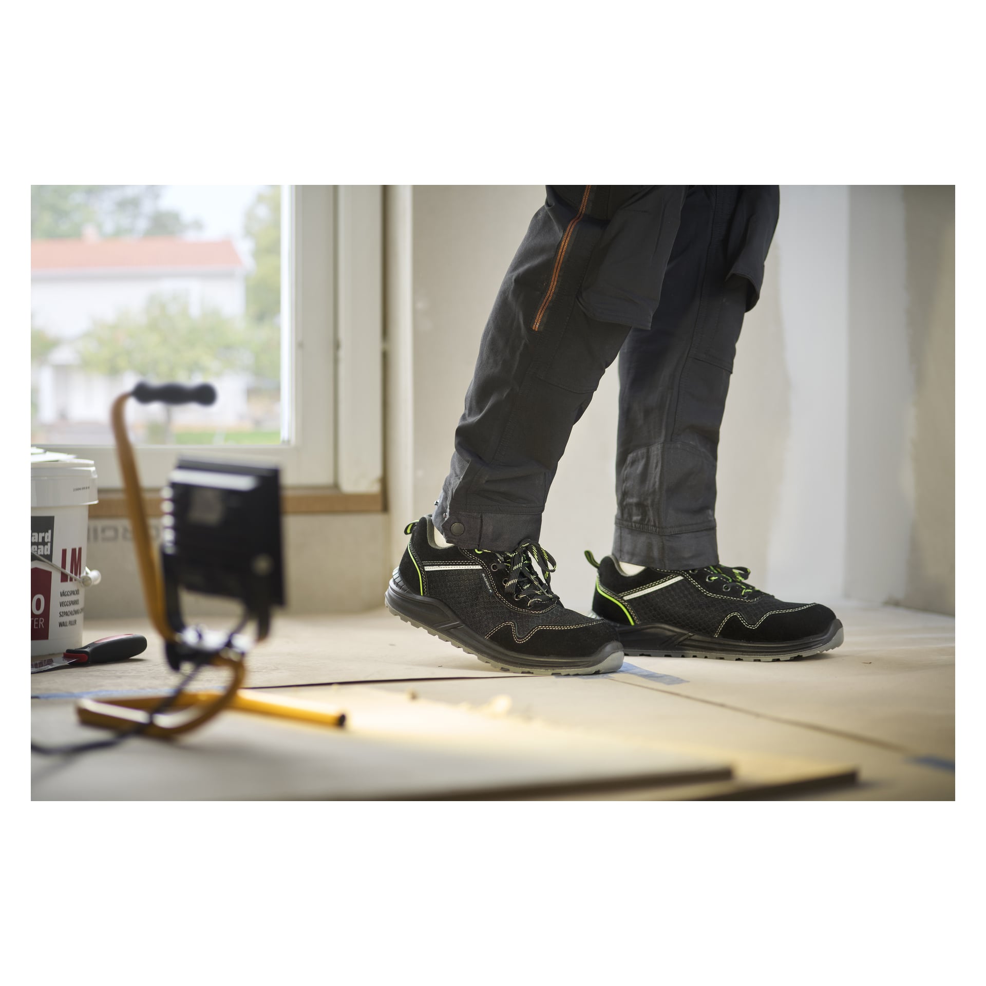 Ktoś w Bluewear Safety Shoes Toledo i ciemnych spodniach stoi na ochronnej podłodze, obok z lampką roboczą i wiaderkiem z farbą.
