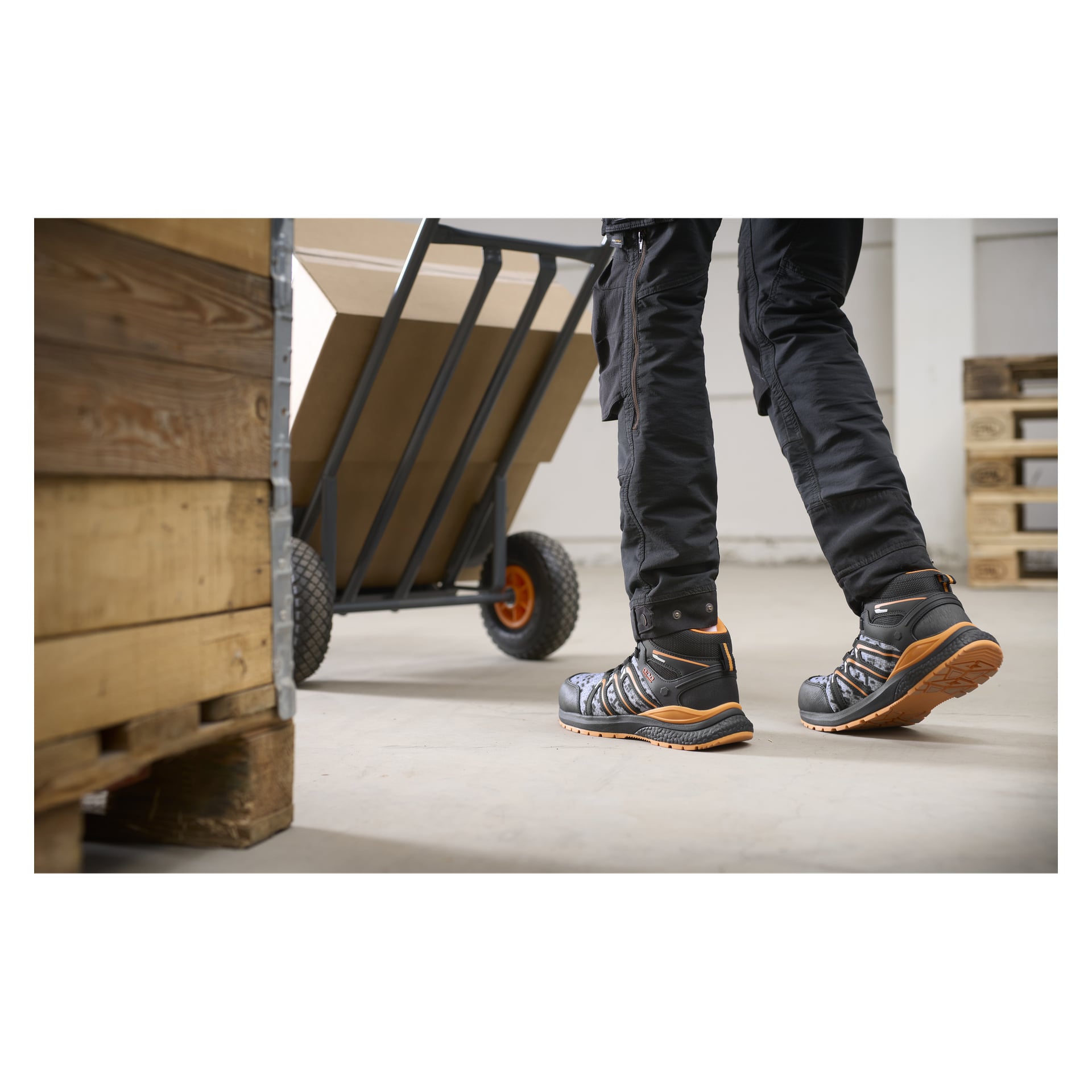 Osoba w czarnych spodniach roboczych i butach Bluewear Pro z szybkim sznurowaniem Phoenix przechodzi obok palety i wózka ręcznego z dużym pudełkiem w środku.